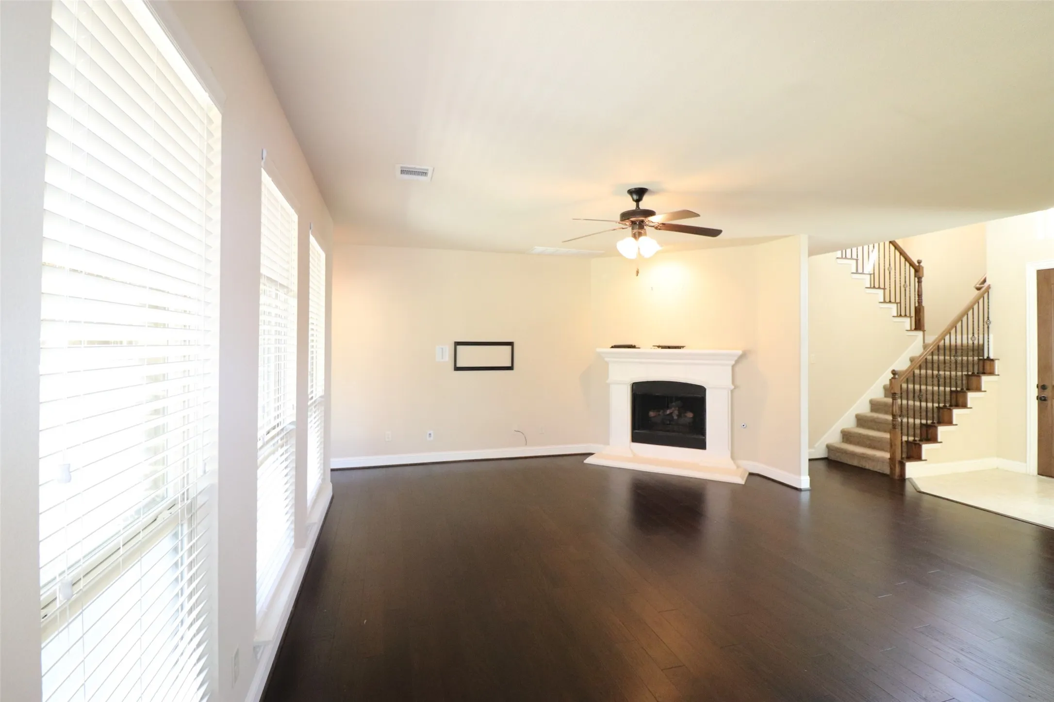 Spacious room featuring wood-finish flooring, multiple windows with blinds, a white mantel fireplace, and a ceiling fan