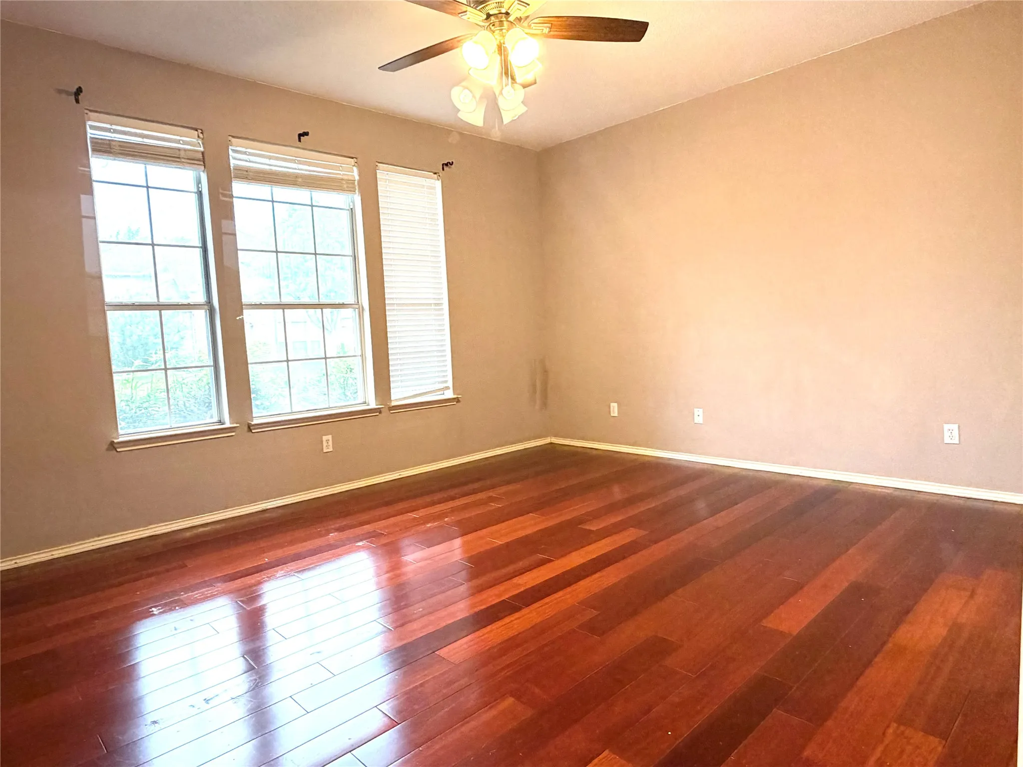 Spacious room featuring rich wood-finish flooring, three large windows with blinds, and a ceiling fan with integrated lighting