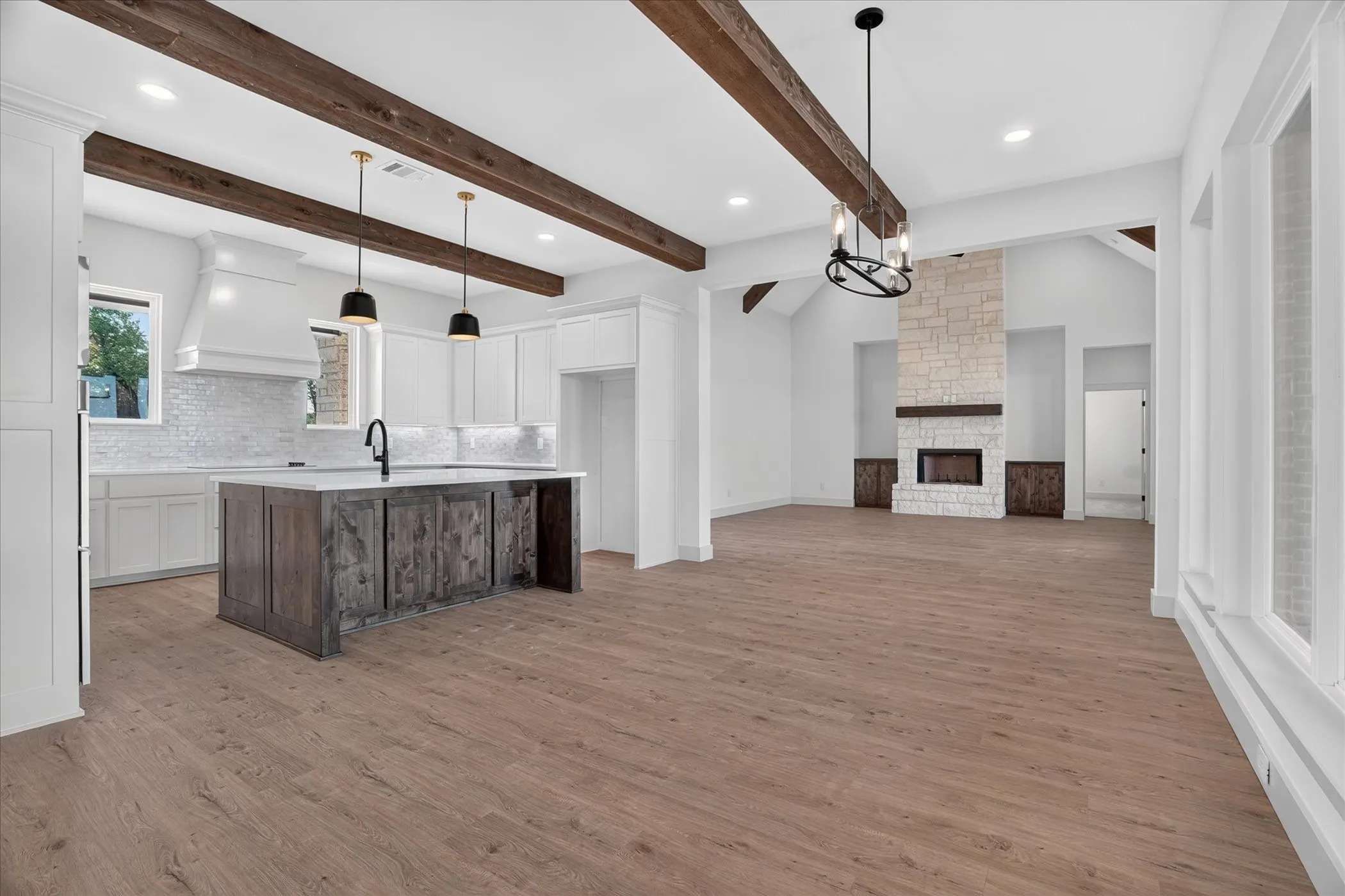 Open-concept living space featuring wood-finish flooring, exposed wood beams, a stone fireplace, and a kitchen island with built-in cabinetry