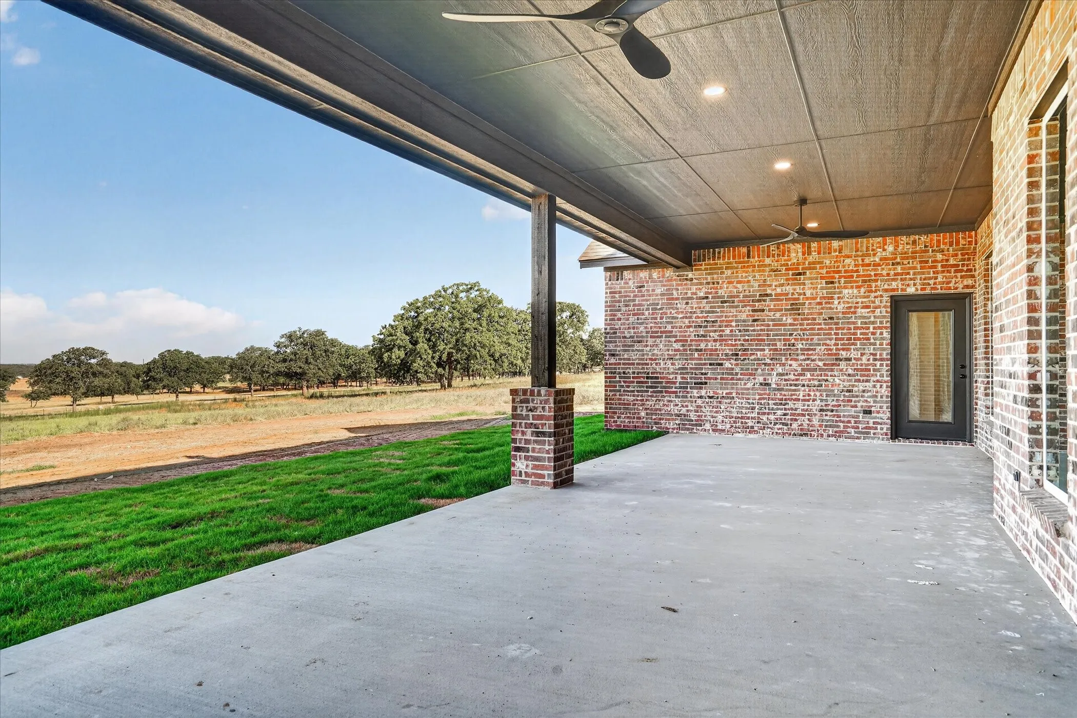 Expansive covered patio with concrete flooring, brick exterior, ceiling fans, and recessed lighting overlooking a landscaped lawn and mature trees