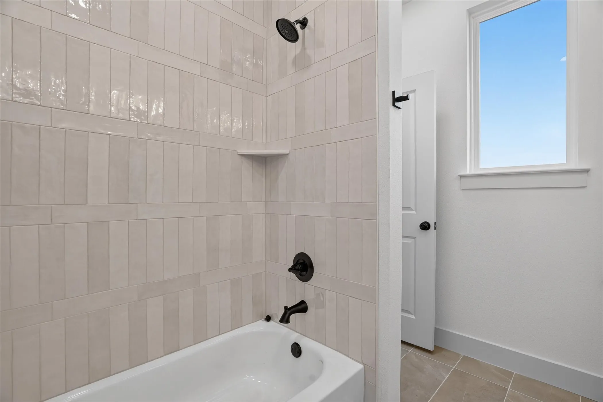 Bathtub with matte black fixtures, tile surround with integrated shelving, and a bright window
