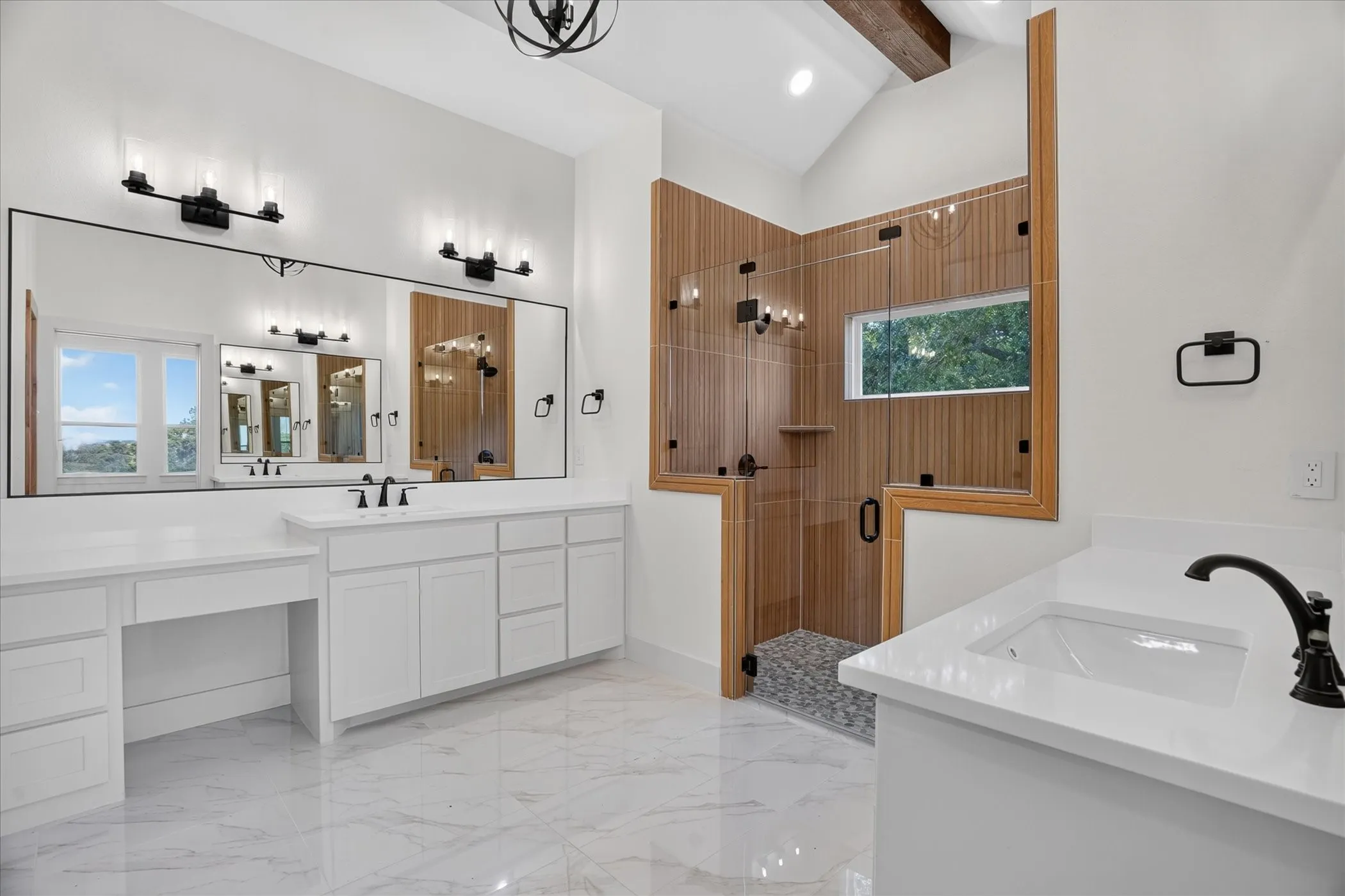Vaulted ceiling with exposed beam, white cabinetry with built-in vanity, wood-look tiled shower with glass enclosure, white rectangular under-mount sinks, and large frameless mirror