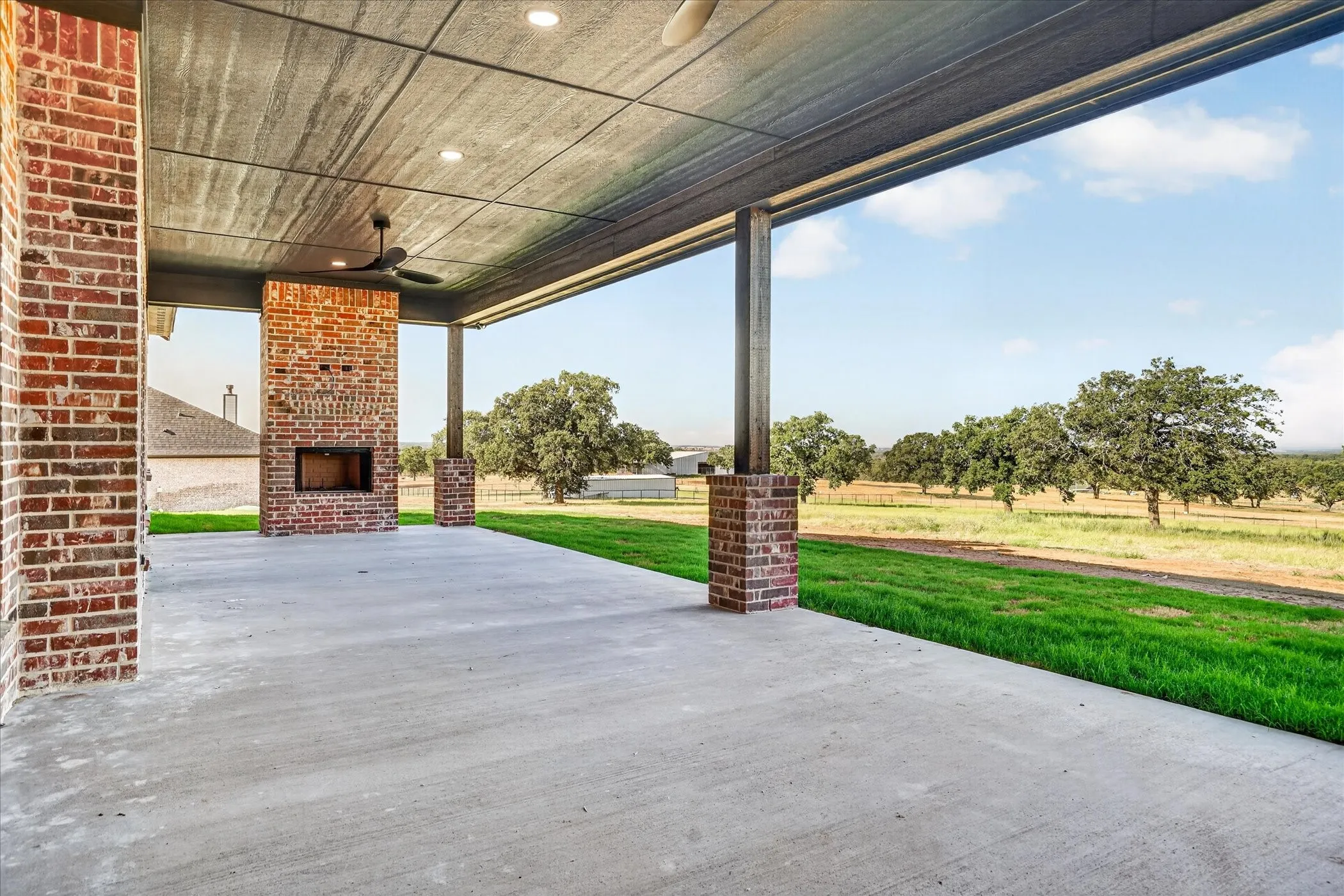 Expansive covered patio featuring a brick-clad outdoor fireplace, exposed brick columns, and a ceiling fan with integrated lighting
