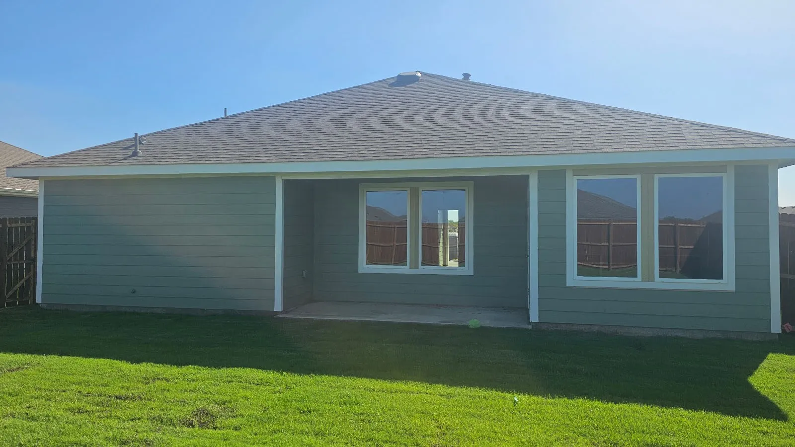 Expansive green lawn, covered concrete patio, multiple large windows, horizontal siding exterior, and dark shingle roof