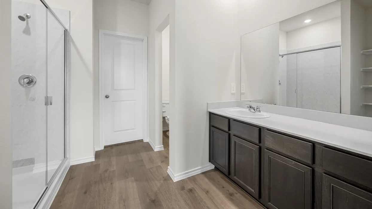 Wood-finish flooring with a glass-enclosed shower, dark wood vanity with a white countertop, and expansive wall-mounted mirror