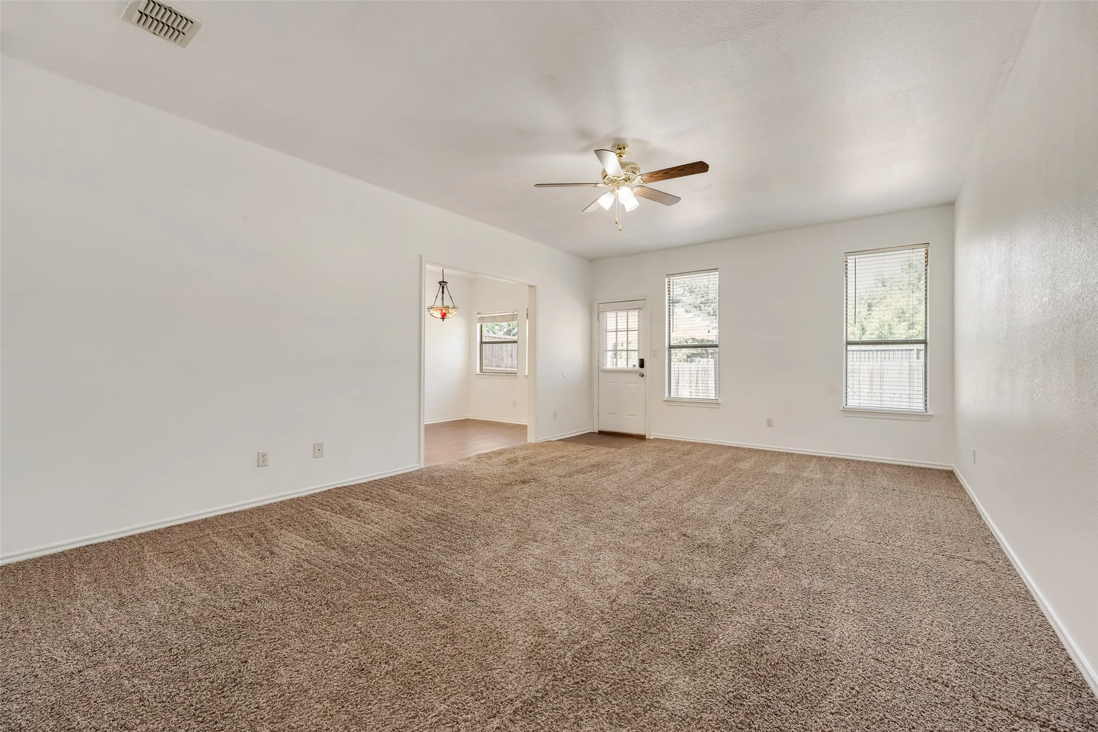 Spacious living area with carpet flooring, a ceiling fan, a white door with divided lites, and two windows with blinds