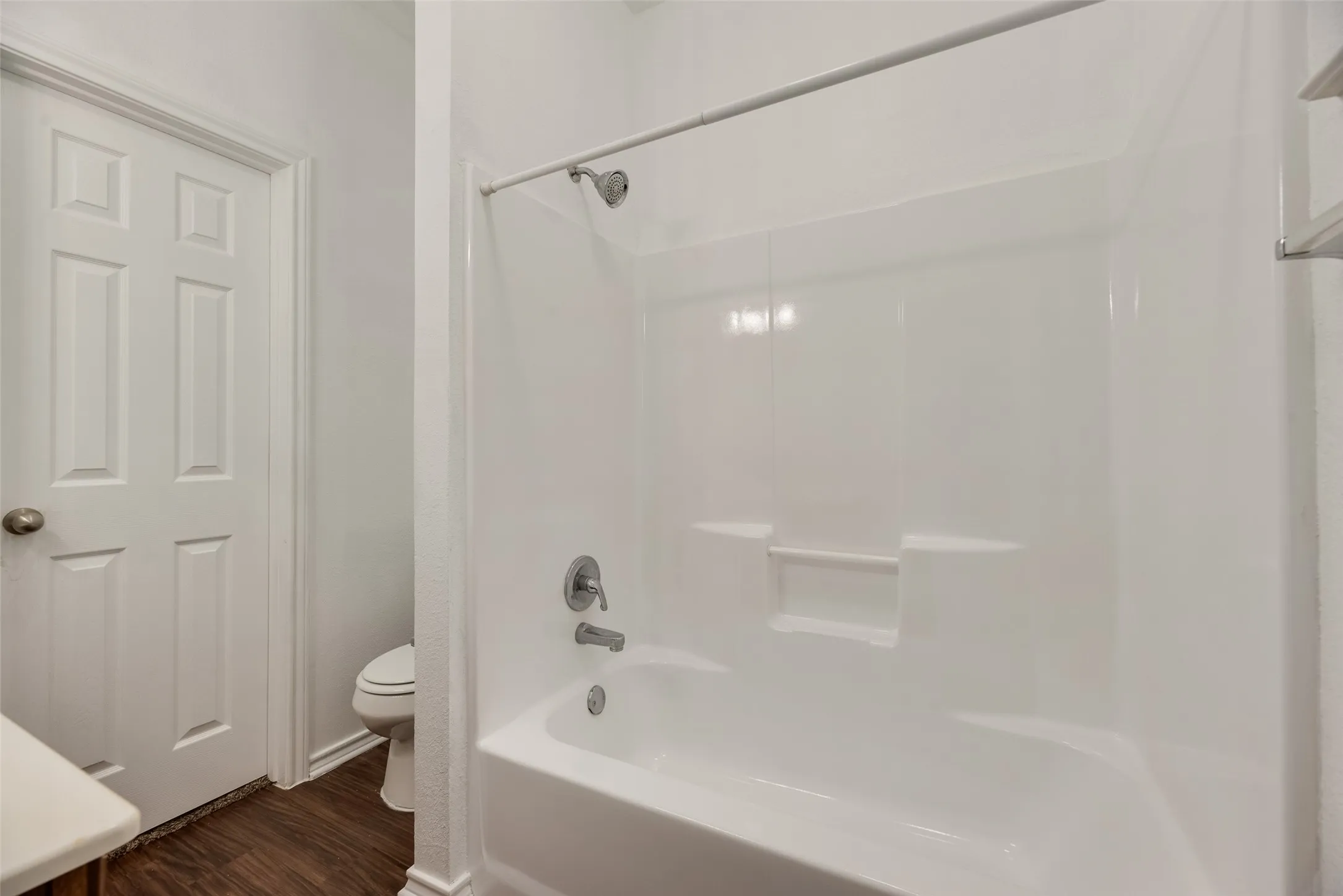 White paneled door, shower/tub combination, integrated shower shelving, chrome shower fixtures, and wood-finish flooring
