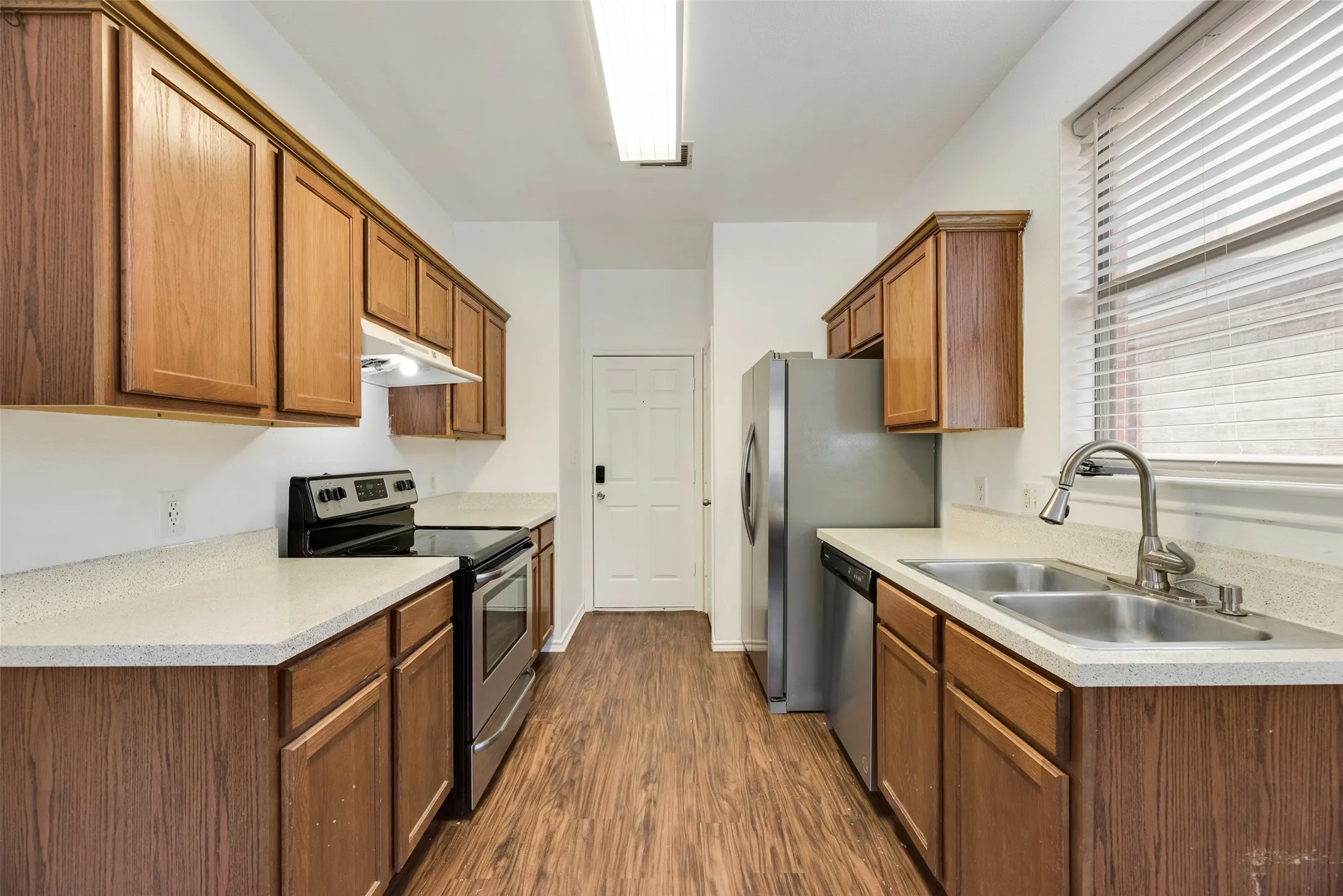 Galley kitchen featuring wood-finish cabinetry, light-toned countertops, stainless steel appliances, and wood-finish flooring