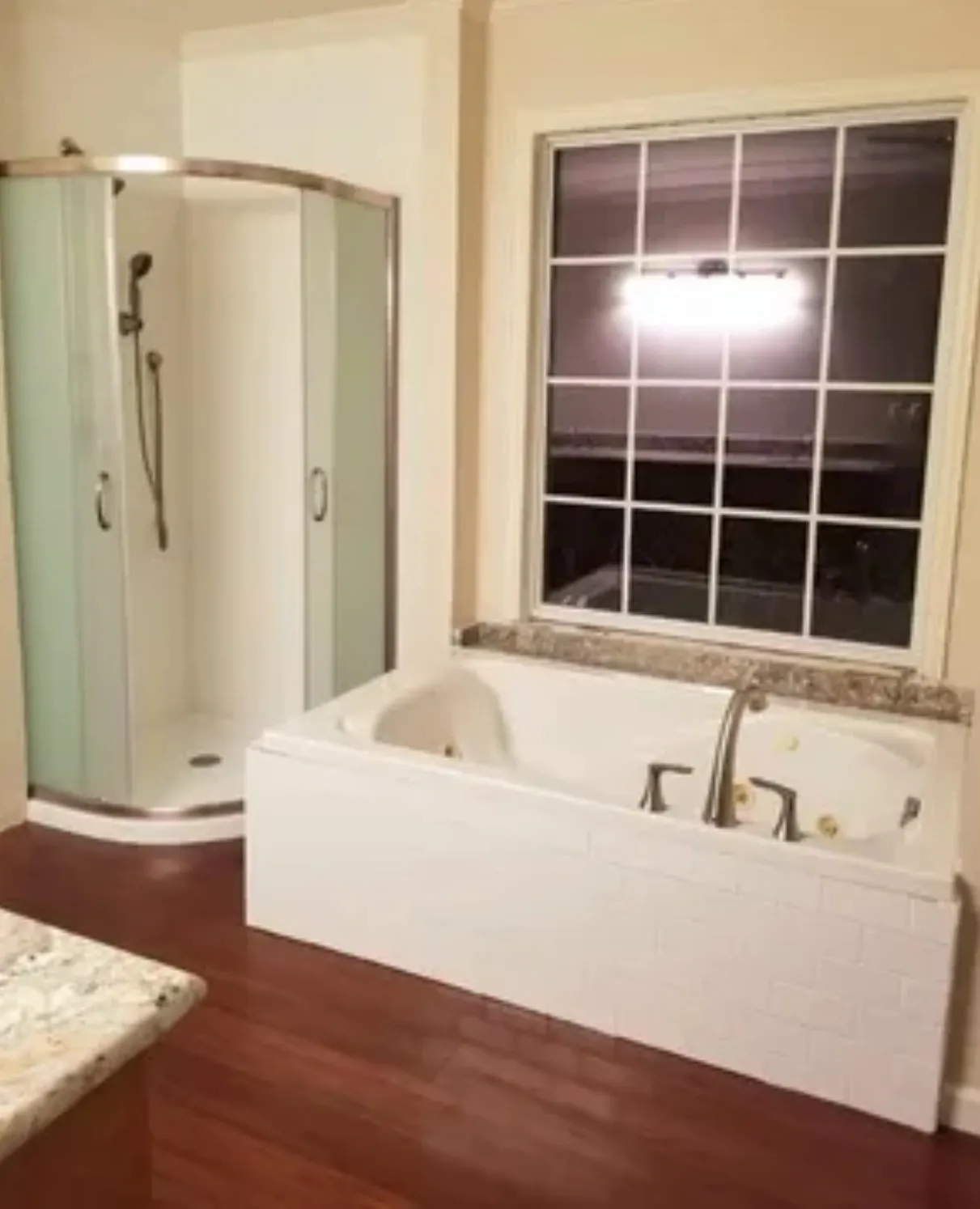 Soaking tub with tiled surround, separate corner shower, wood-finish flooring, and a large grid-pattern window