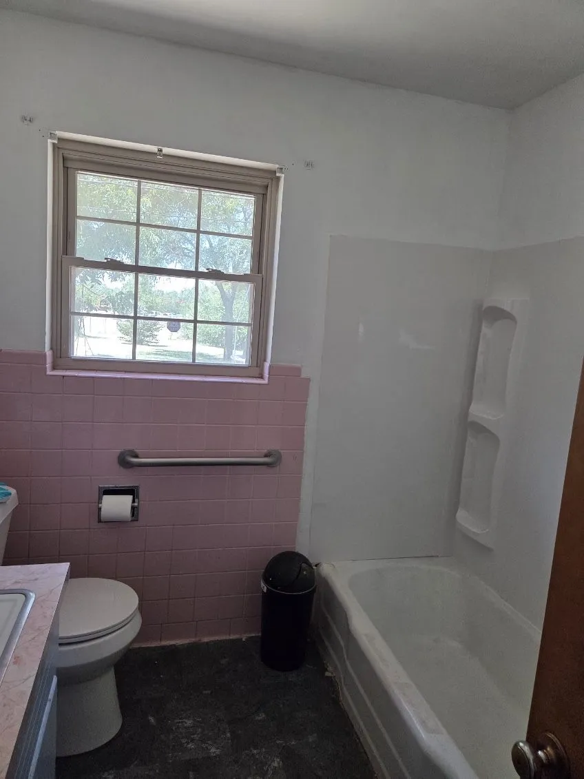 Window with gridded panes, pink tiled wainscoting, wall-mounted grab bar, and built-in bathtub