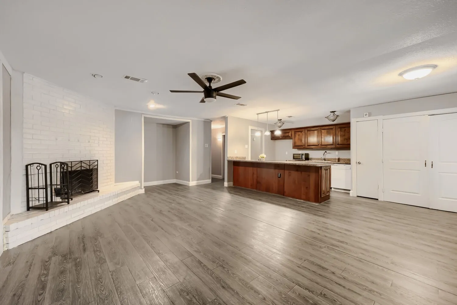 Open-concept living area featuring a white brick fireplace, wood-finish flooring, and a kitchen island with pendant lighting
