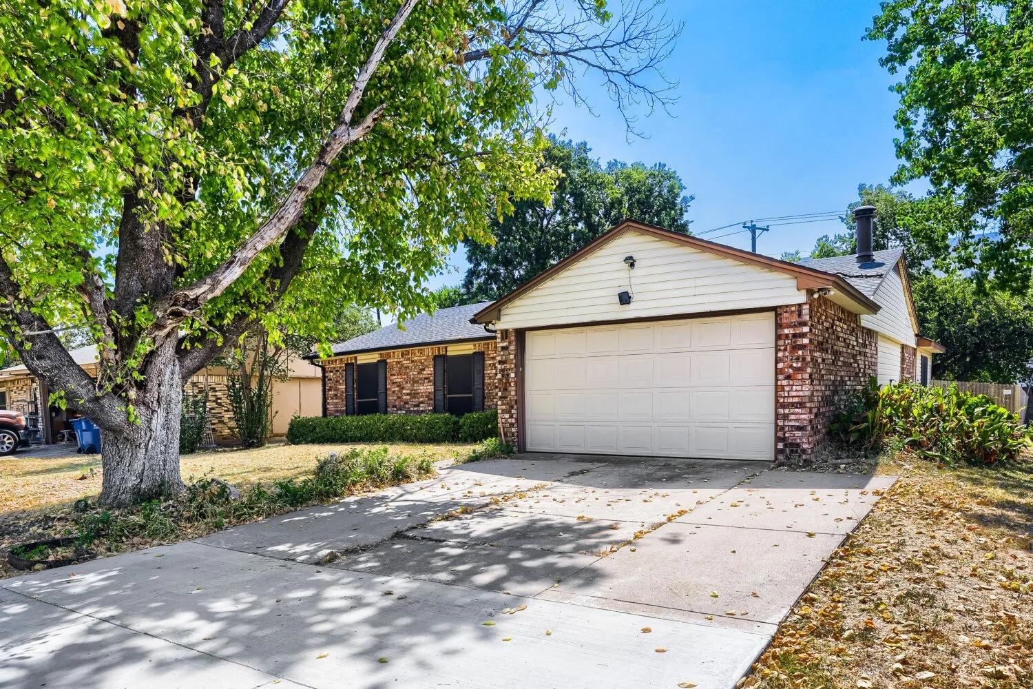 Charming brick home featuring a spacious two-car garage with a light-colored overhead door, concrete driveway, and mature shade trees framing the front elevation