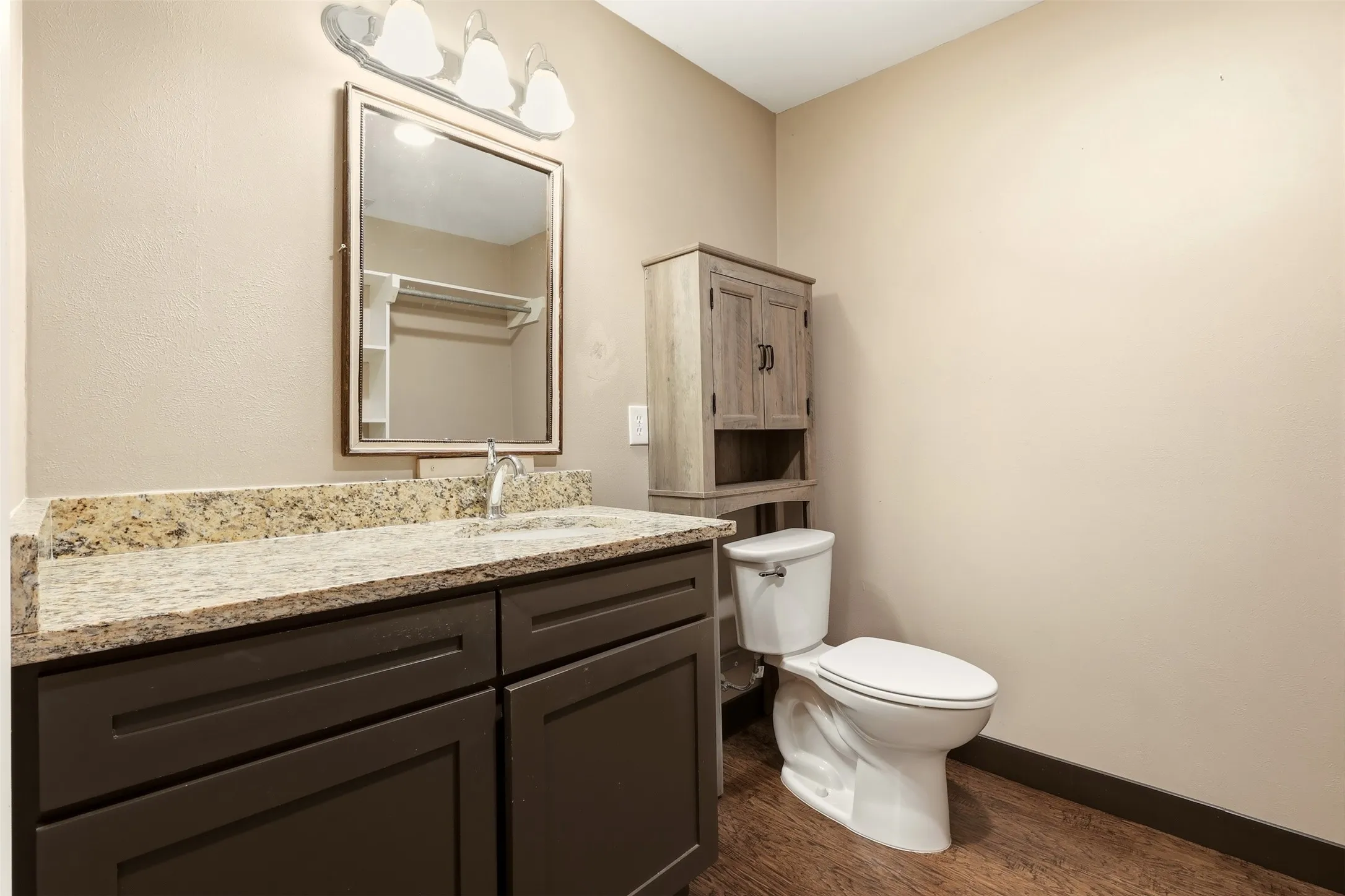 Granite-finish countertop, dark wood-finish vanity, wood-finish flooring, and wall-mounted vanity lighting
