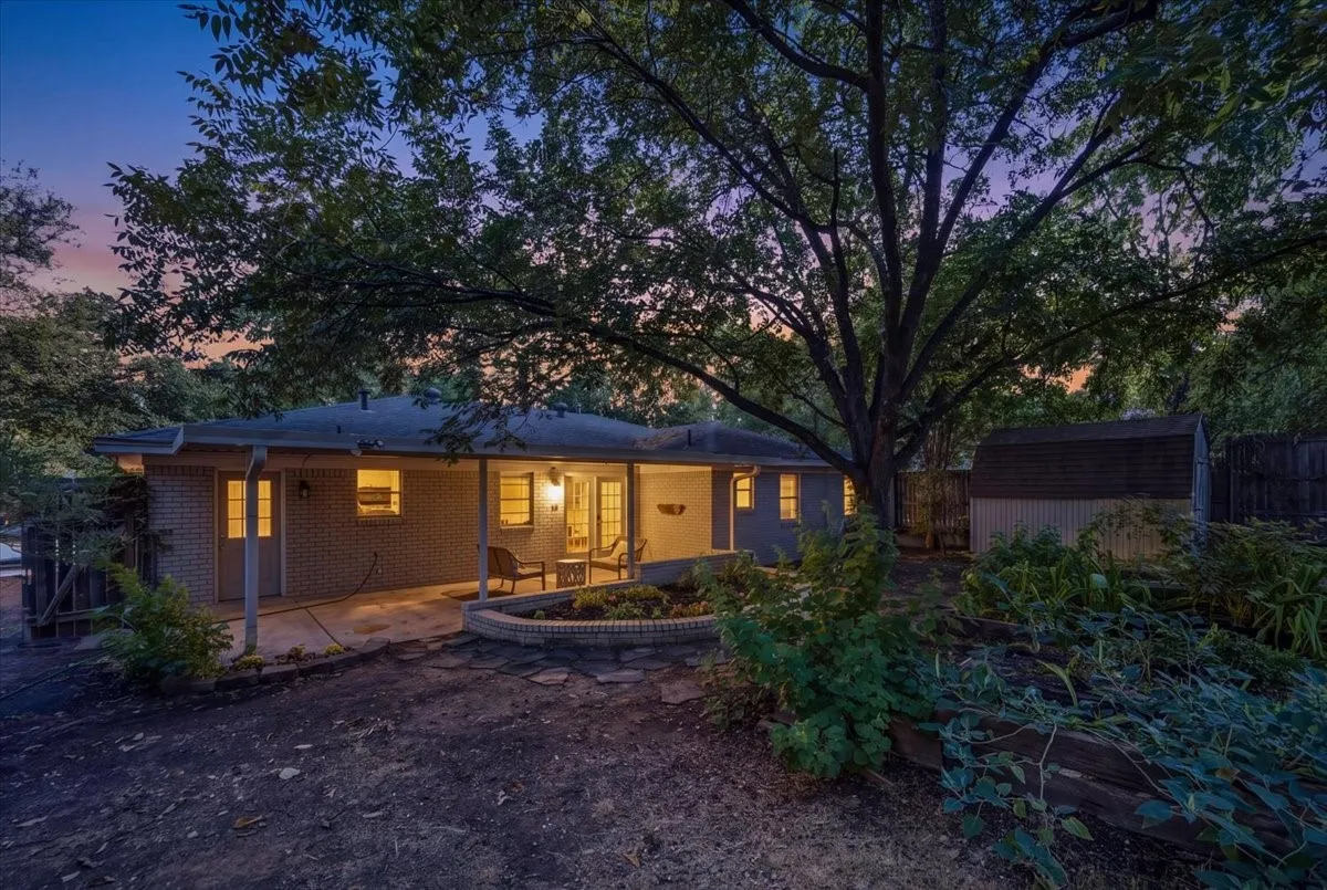 Modern residence bathed in warm ambient lighting, featuring a clean-lined exterior with large windows, a covered front porch, and mature shade trees framing the property