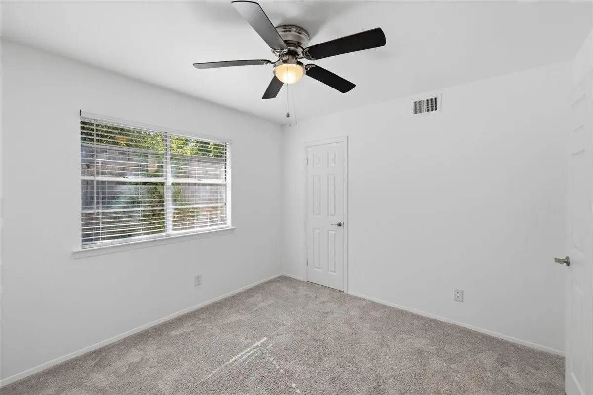 Plush gray carpeting, white painted walls, multi-pane window with blinds, paneled door, and a ceiling fan with integrated lighting