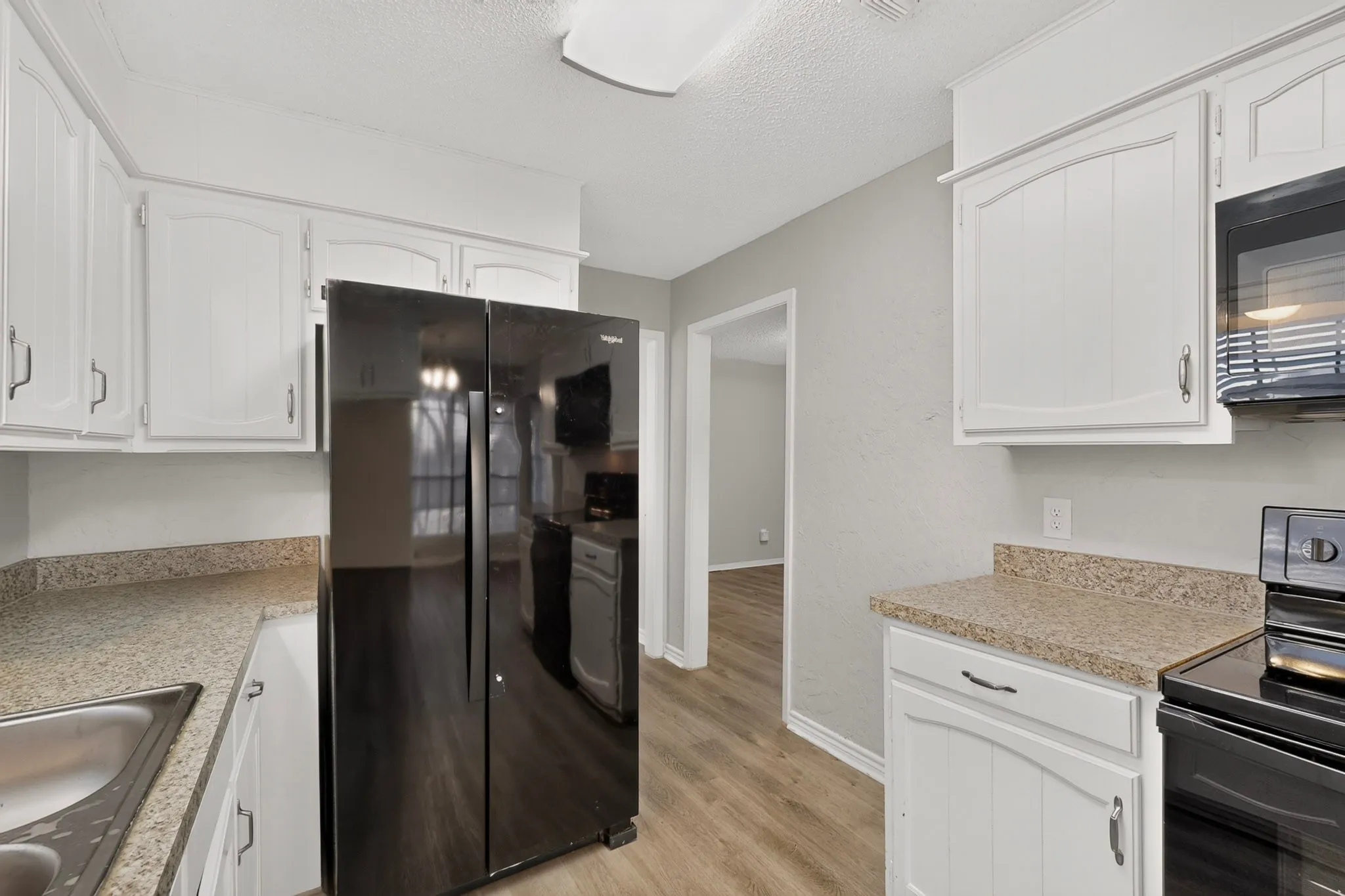 White cabinetry with black hardware, black side-by-side refrigerator, under-mount stainless steel sink, black built-in microwave, and wood-finish flooring