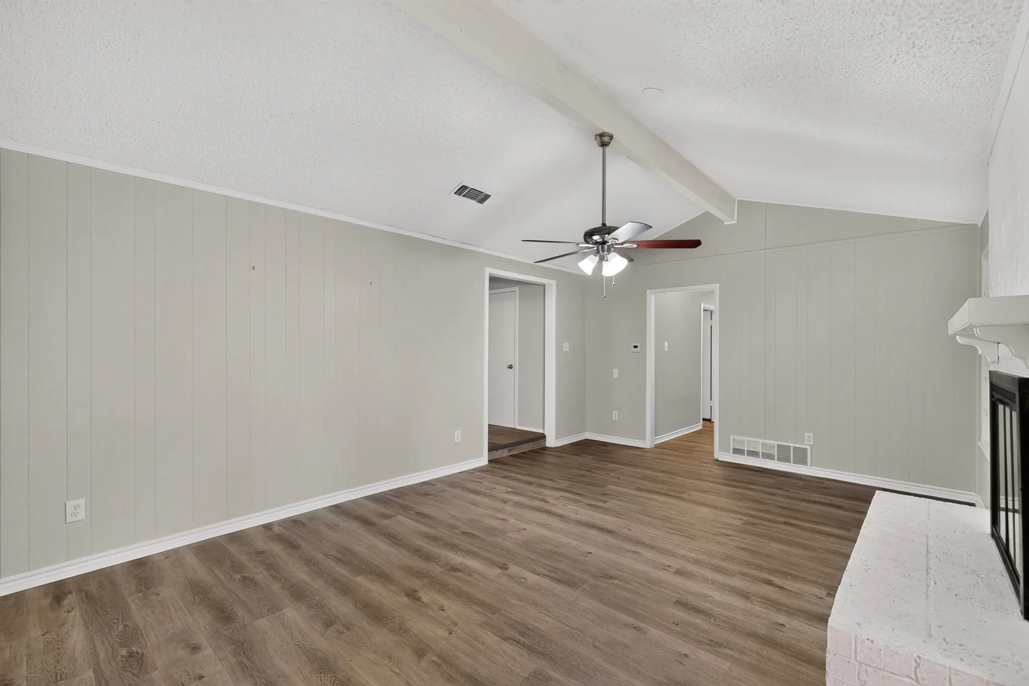 Vaulted ceiling with exposed beam, wood-finish flooring, brick fireplace with mantel, ceiling fan with integrated lighting, and vertical paneling on walls