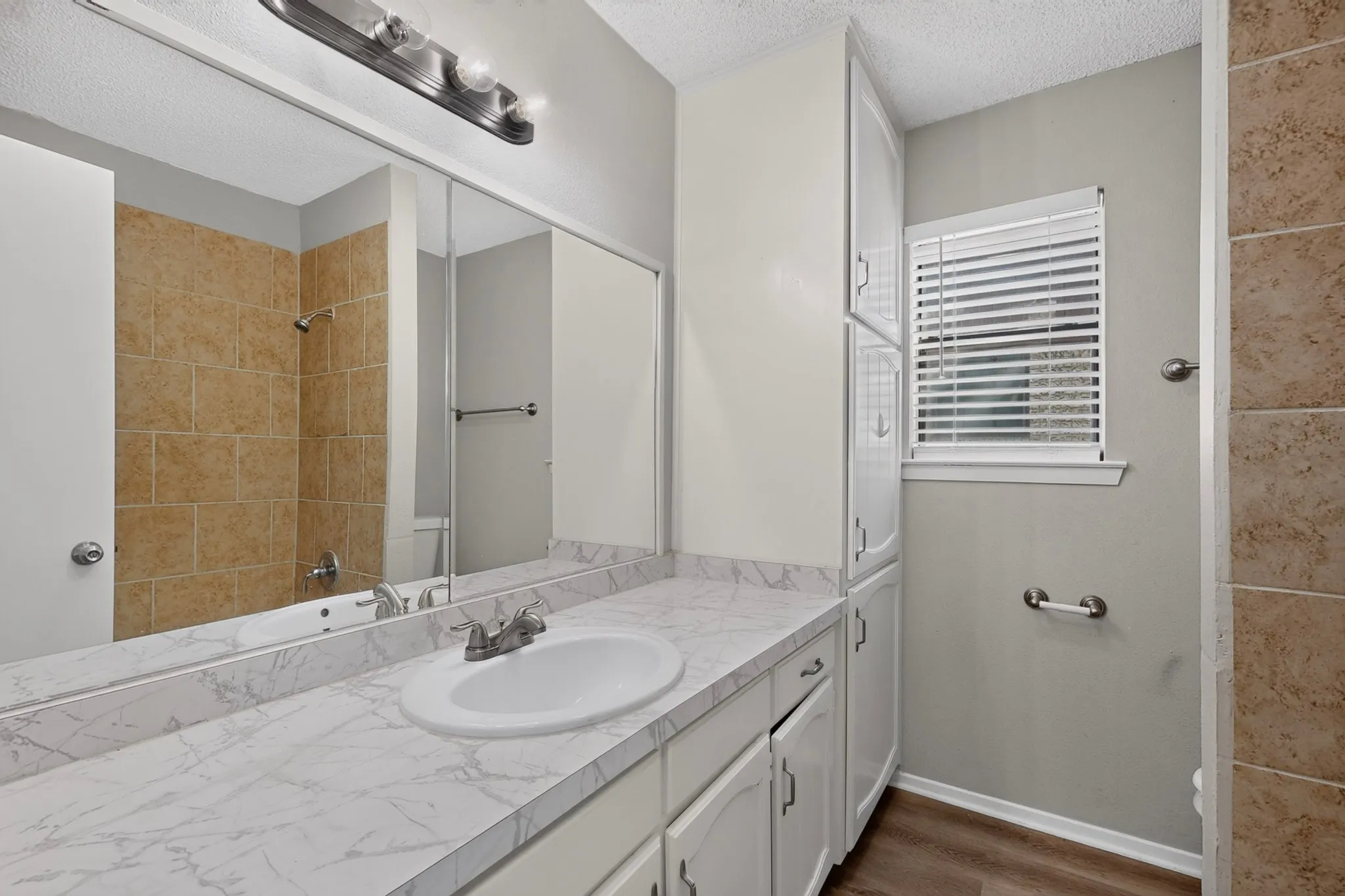 Full-width vanity mirror, spacious countertop with integrated sink, wood-finish flooring, tiled shower surround, and built-in storage cabinetry