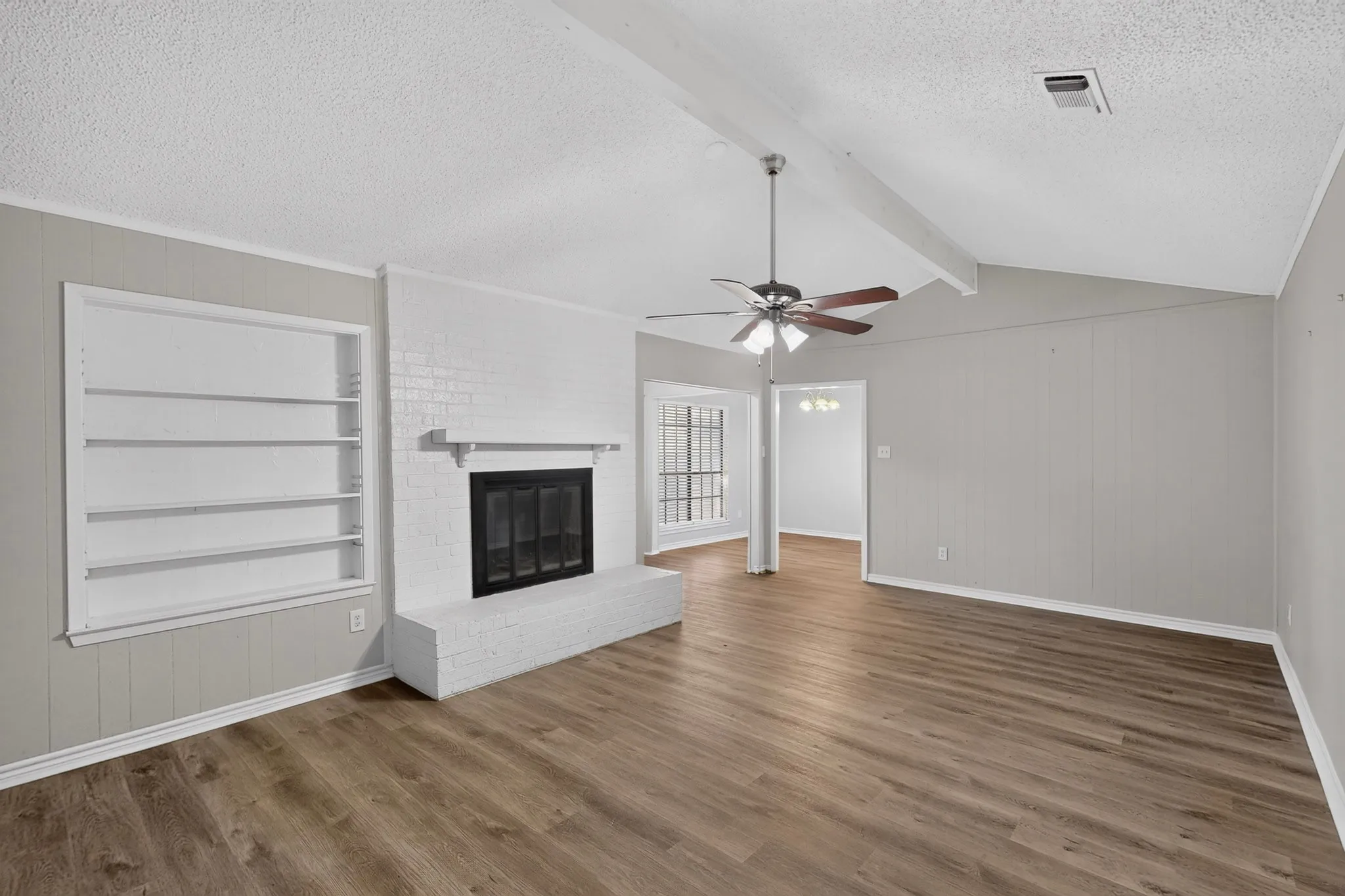Vaulted ceiling with exposed beams, wood-finish flooring, white brick fireplace, built-in shelving, and ceiling fan