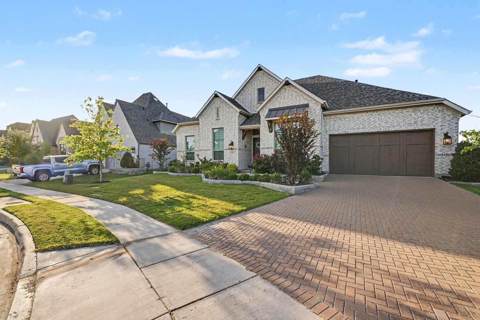 Brick exterior with dark roof shingles, brick paver driveway, manicured landscaping, and an attached garage