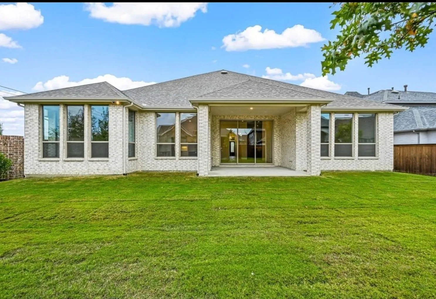 Expansive green lawn, light-colored brick exterior, covered patio, multiple large windows, and sliding glass doors