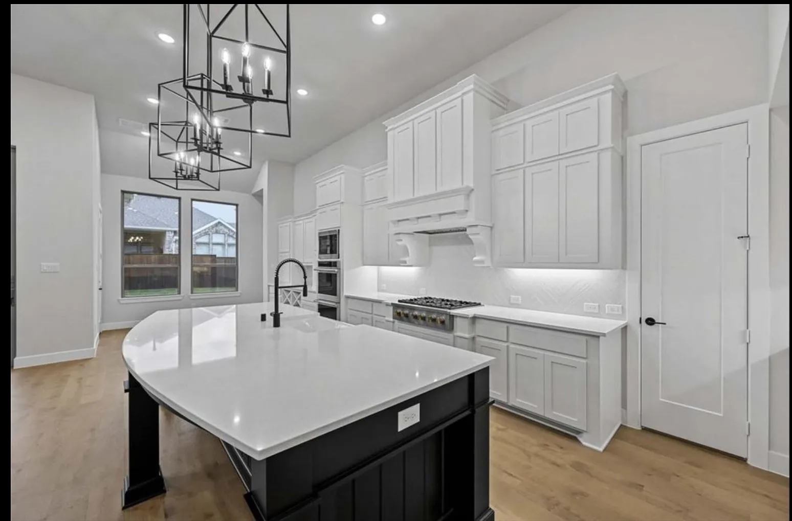Open-concept kitchen featuring a curved island with a light-toned countertop, black-finish pendant lighting, light wood-finish flooring, and white cabinetry