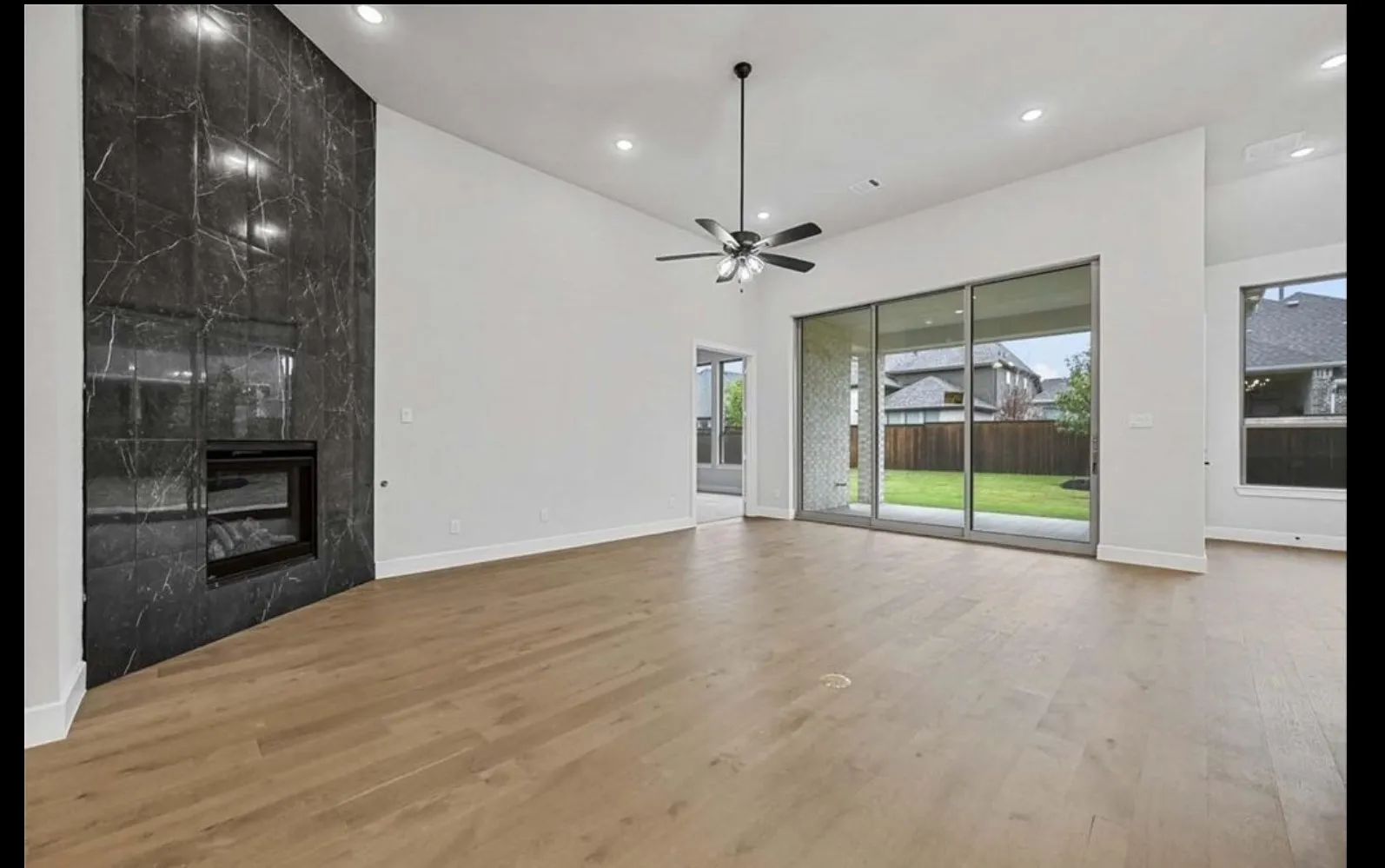 Spacious open-concept living area featuring warm wood-finish flooring, a striking dark stone fireplace with integrated fireplace insert, ceiling fan with brushed metal finish, and sliding glass doors opening to the backyard