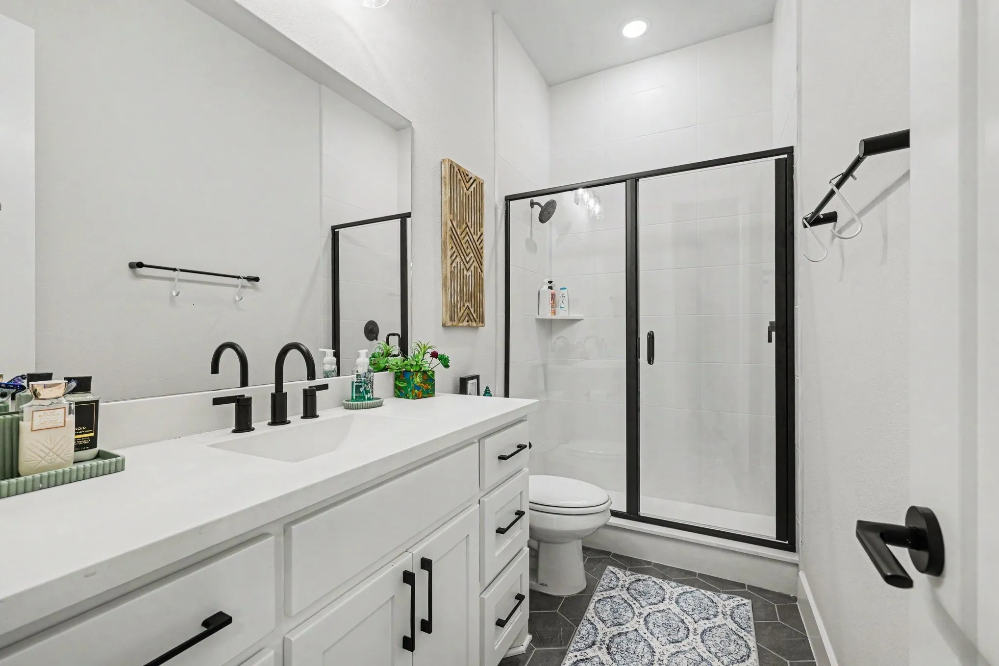 White vanity with integrated sink, black matte fixtures, frameless mirror, glass-enclosed shower, and hexagonal floor tiling