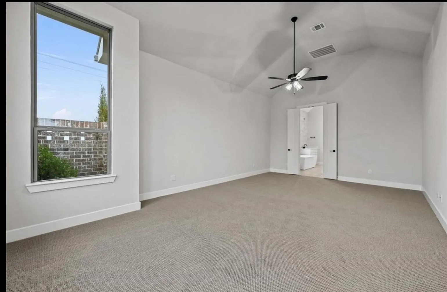 Vaulted ceiling with a ceiling fan, neutral wall paint, light-colored carpet flooring, a tall rectangular window, and double doors opening to a bathroom with a freestanding tub
