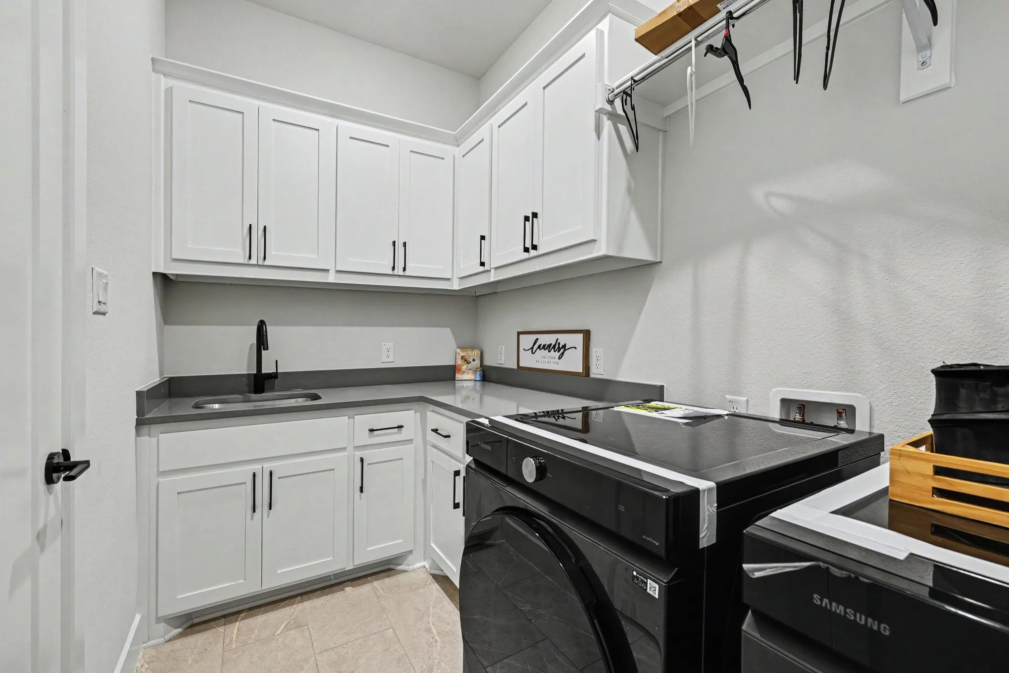 White cabinetry with matte black hardware, dark gray countertops, stainless steel sink with a matte black faucet, and light-toned floor tiling