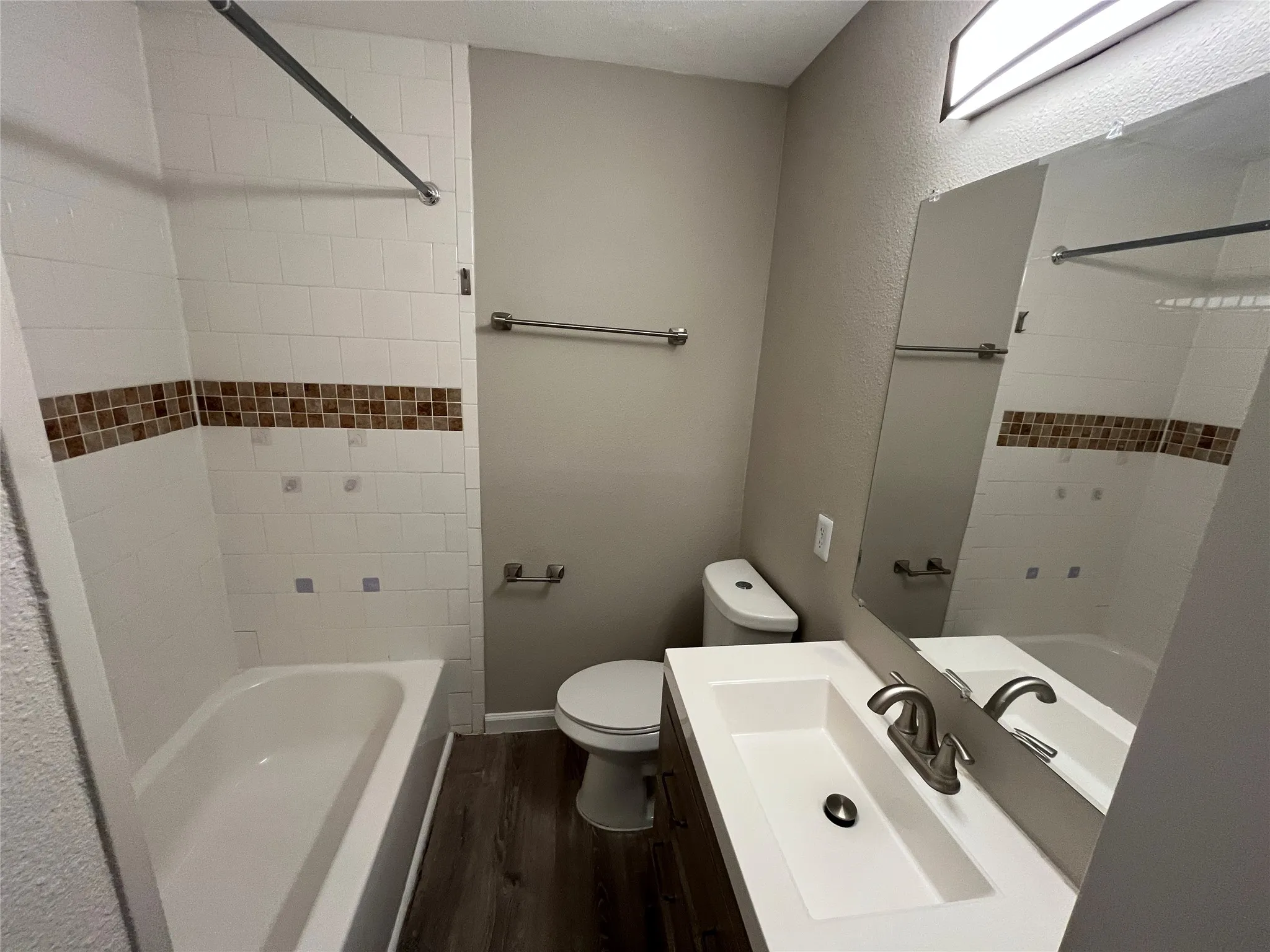 Bathroom featuring wood-finish flooring, a white vanity with an integrated sink, a large wall mirror, and a tub with white tile surround and decorative accents
