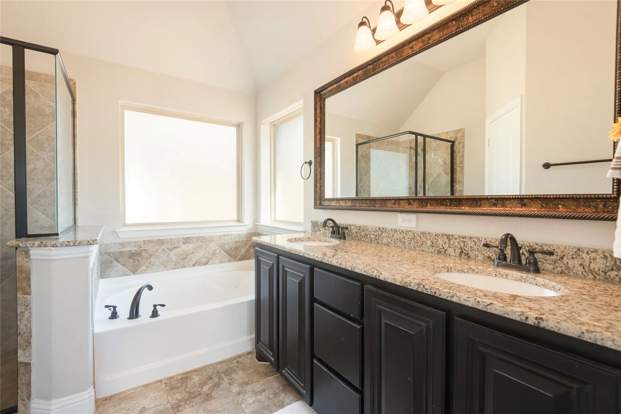 Double vanity with granite countertops, rich dark cabinetry, an oversized framed mirror, and a built-in soaking tub with a tiled surround