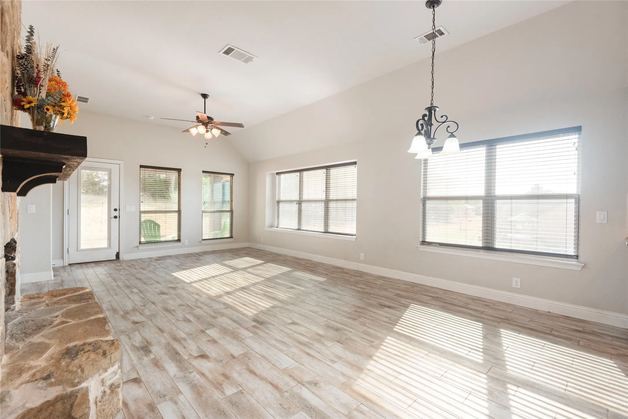Vaulted ceiling with a ceiling fan, stone-clad fireplace with a wood mantle, wood-finish flooring, and multiple large windows with blinds