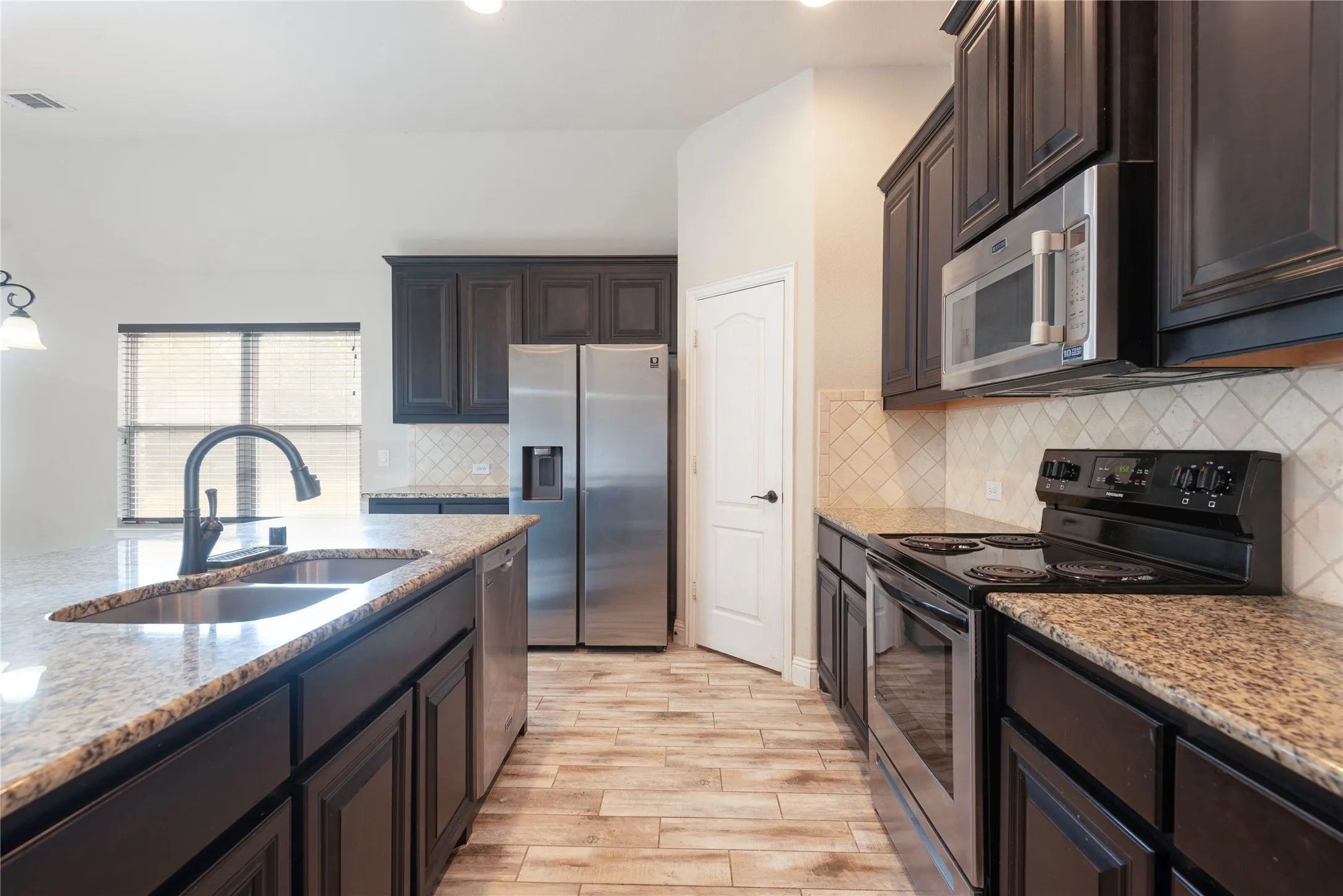 Kitchen featuring dark wood cabinetry, granite countertops, stainless steel appliances, tile backsplash, and wood-look title flooring