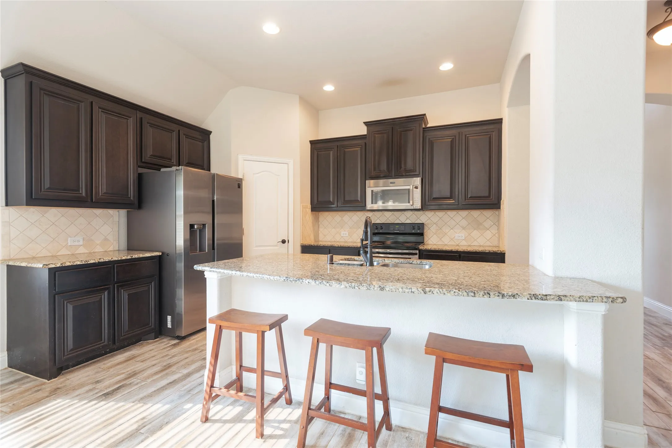 Kitchen featuring dark wood cabinetry, granite countertops, tile backsplash, stainless steel appliances, and wood-like tile flooring