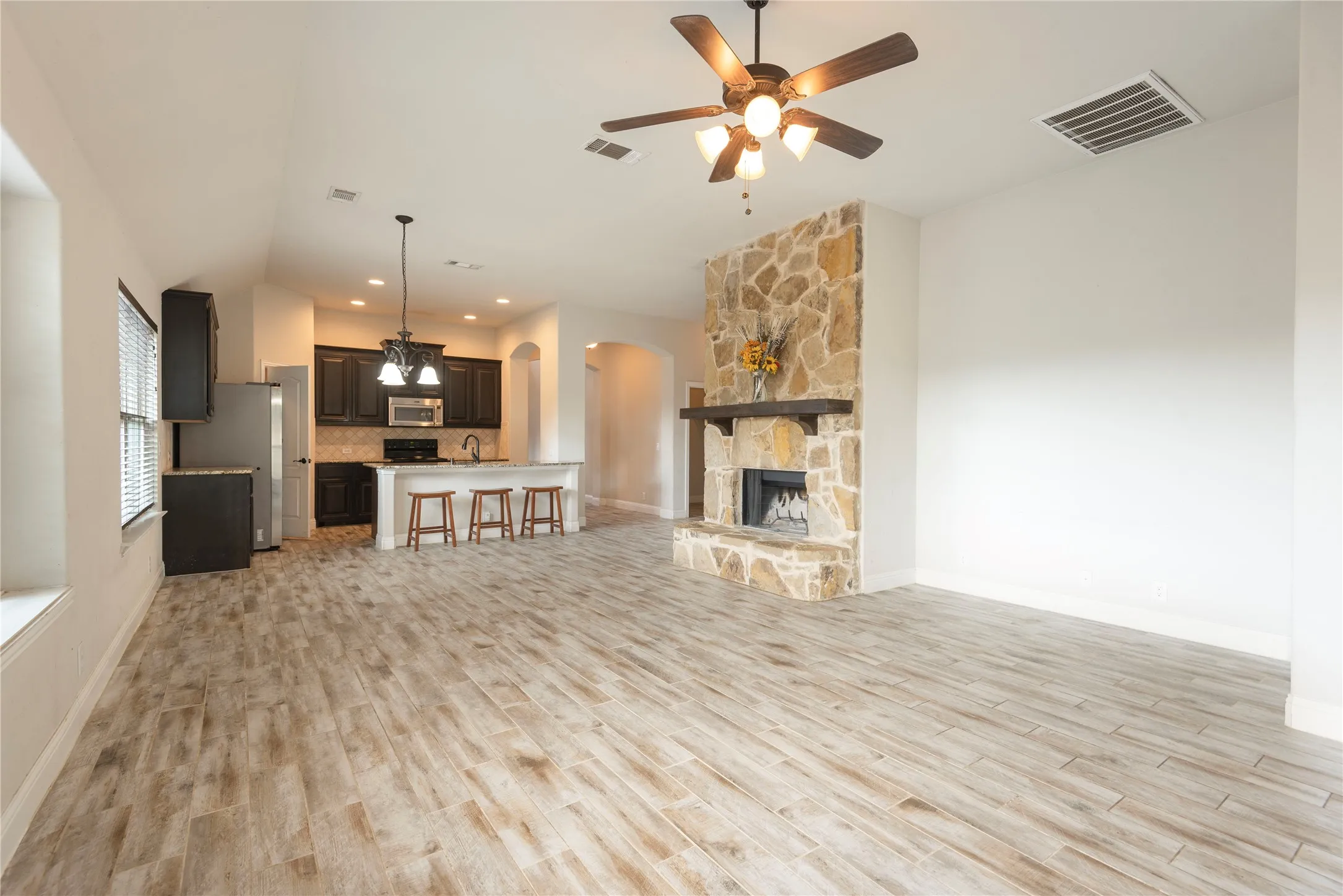 Vaulted ceiling with a ceiling fan, stone-clad fireplace with wood mantle, wood-look title flooring, kitchen island with counter seating, and dark cabinetry