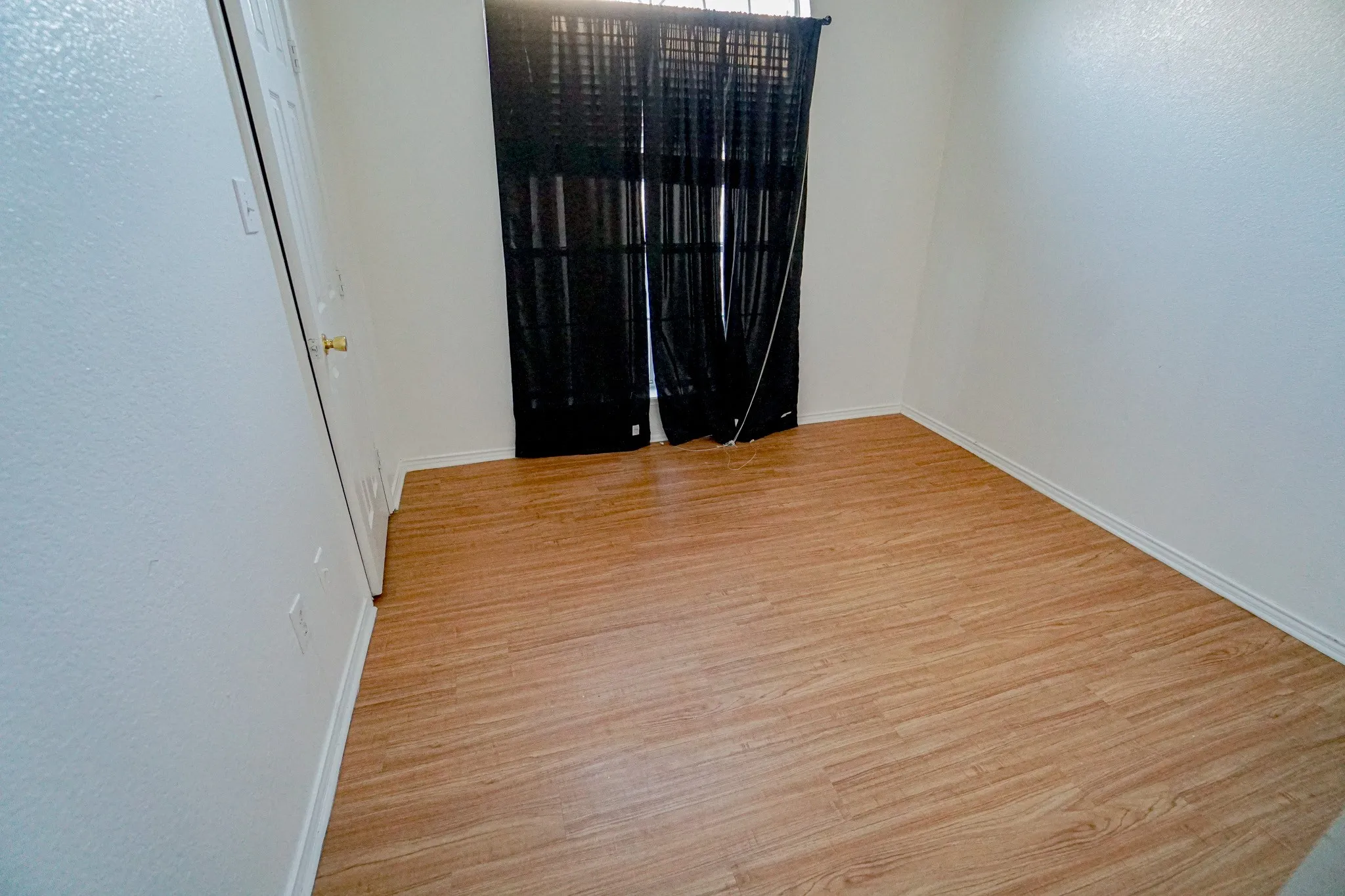 Wood-finish flooring with white baseboards, bright white walls, and a window with dark blinds