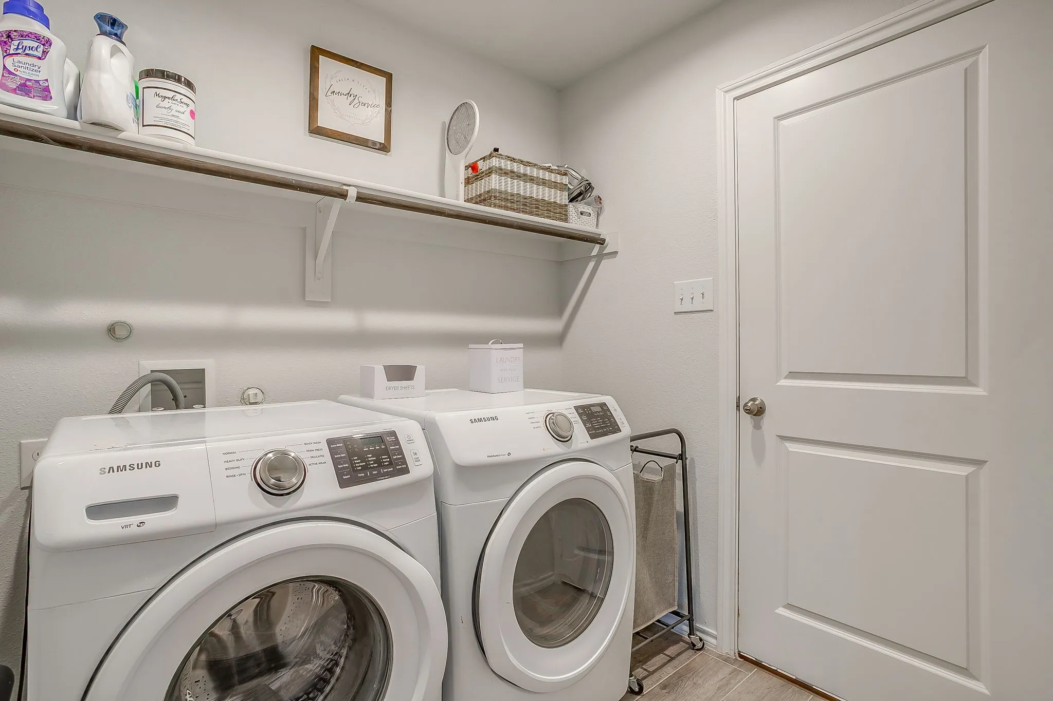 Dedicated laundry room featuring side-by-side Samsung washer and dryer with digital controls, neutral tile flooring, built-in wall shelving with storage baskets, and overhead cabinetry for supplies and linens