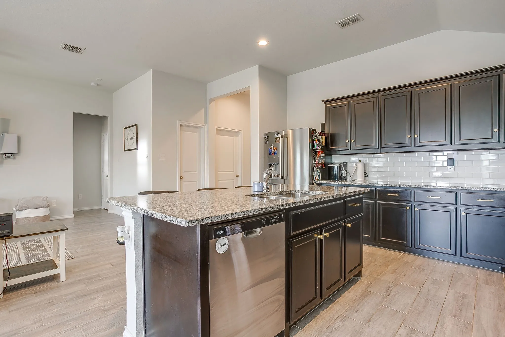 Kitchen featuring a central island with an undermount sink, stainless steel dishwasher, dark wood-finish cabinetry, light-toned countertops, and a white subway tile backsplash