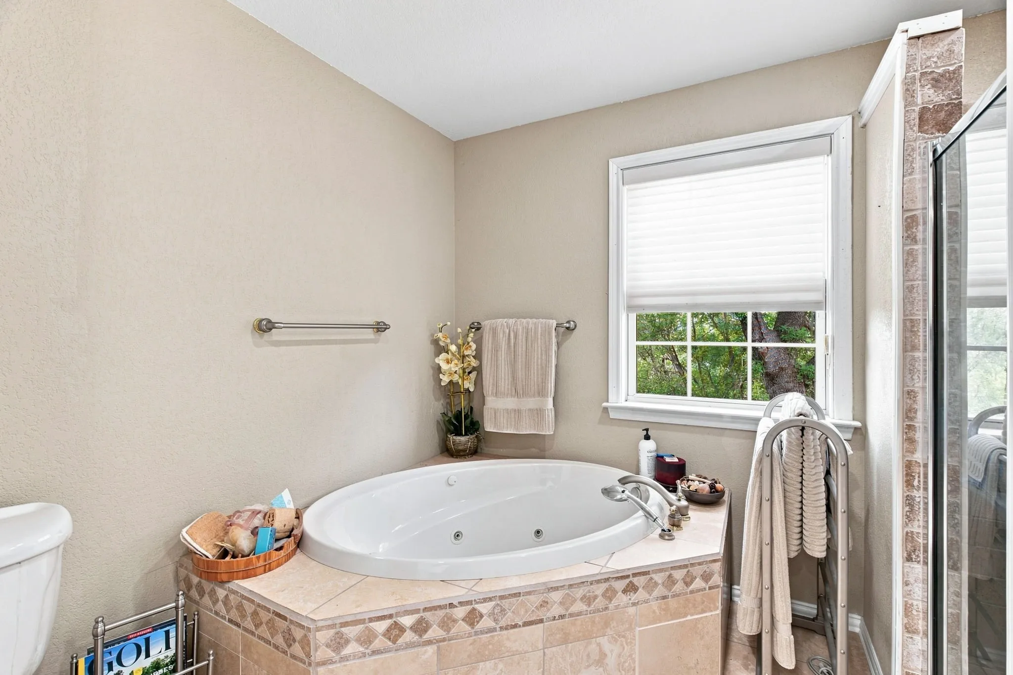 Built-in soaking tub with tile surround, window with privacy blind, and adjacent glass-enclosed shower