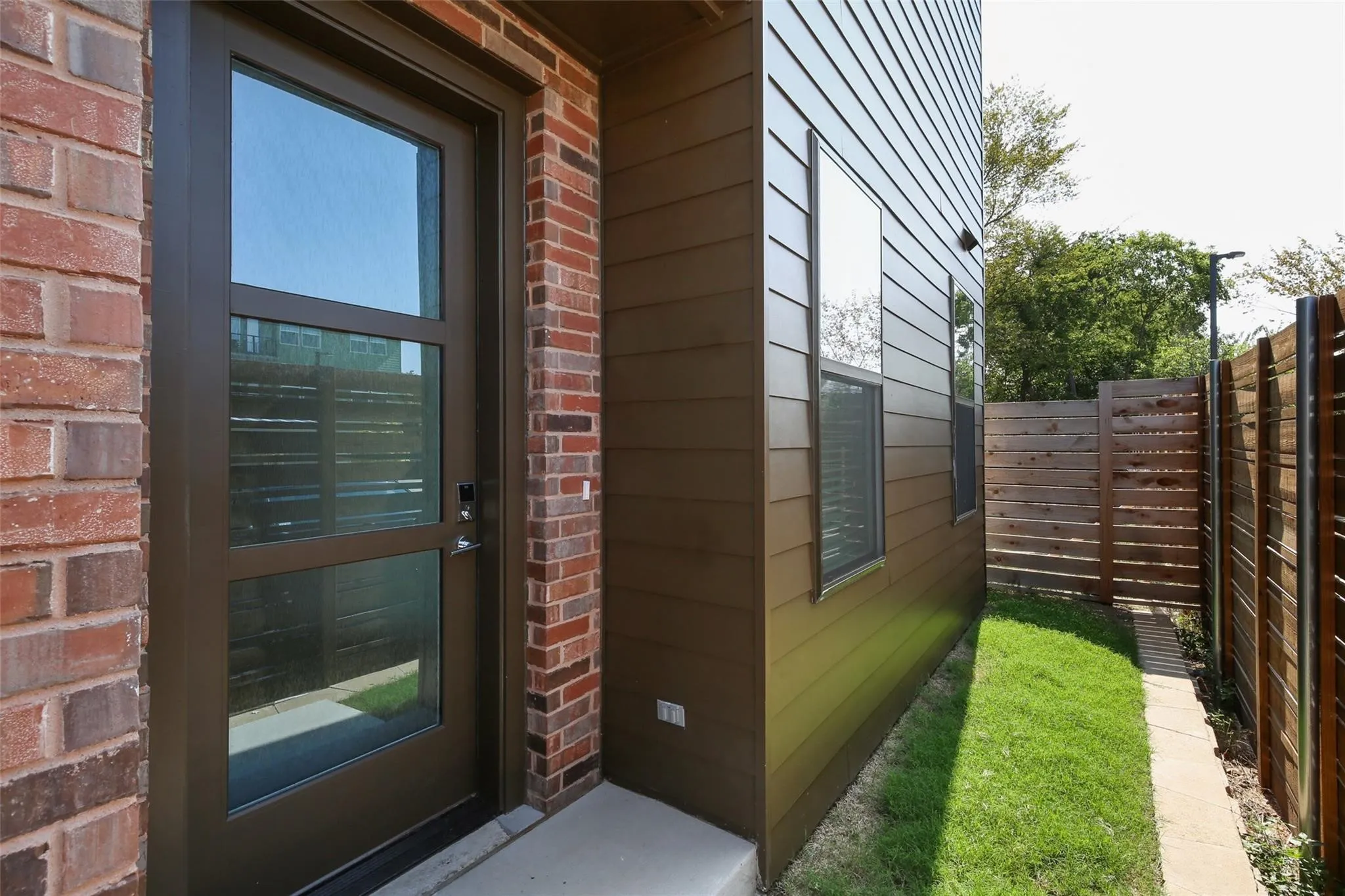 Brick facade, wood-finish siding, modern entry door with glass panels, side yard with green lawn, and wood fencing