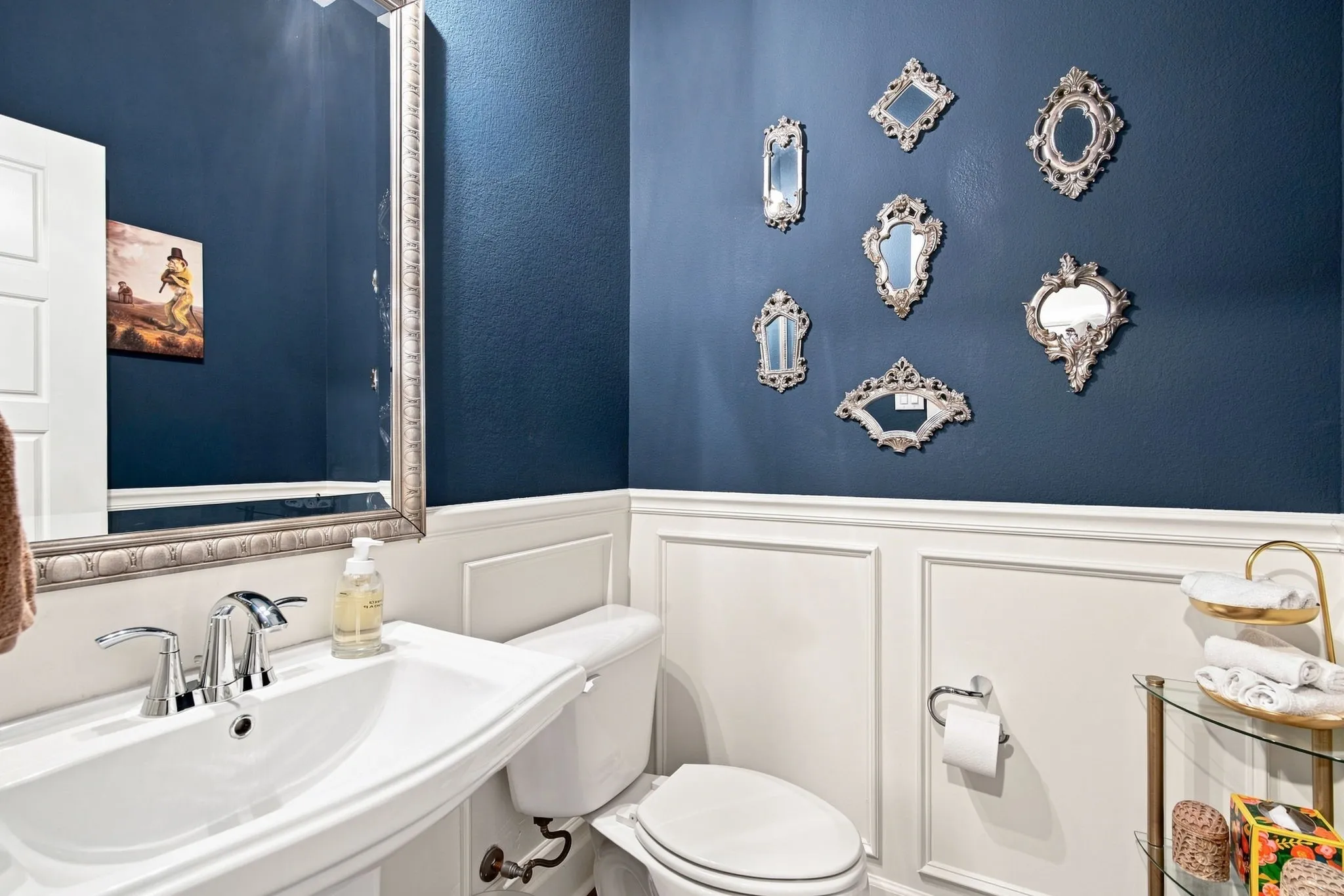 Pedestal sink with chrome faucet, framed mirror, wainscoting, and a wall-mounted toilet paper holder