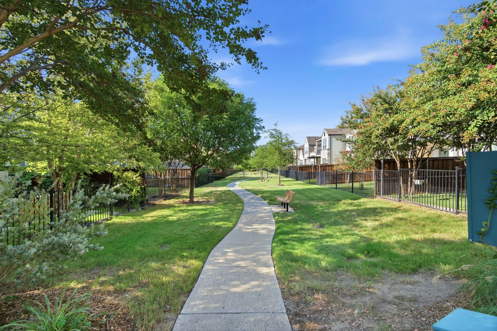 Community green space featuring a paved walkway, mature trees, and perimeter fencing