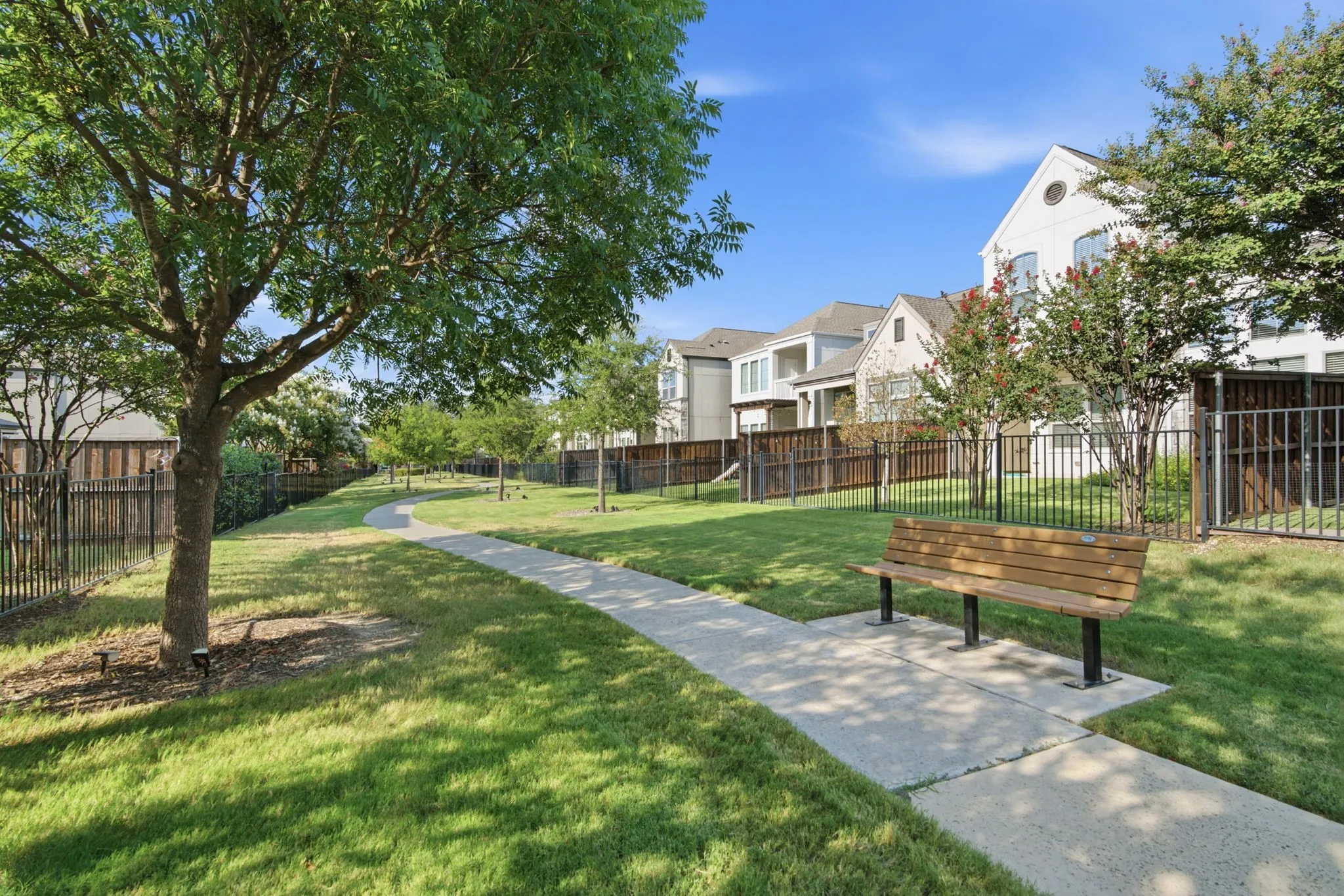 Expansive green space featuring a concrete pathway, mature trees, a wooden bench, and black metal fencing