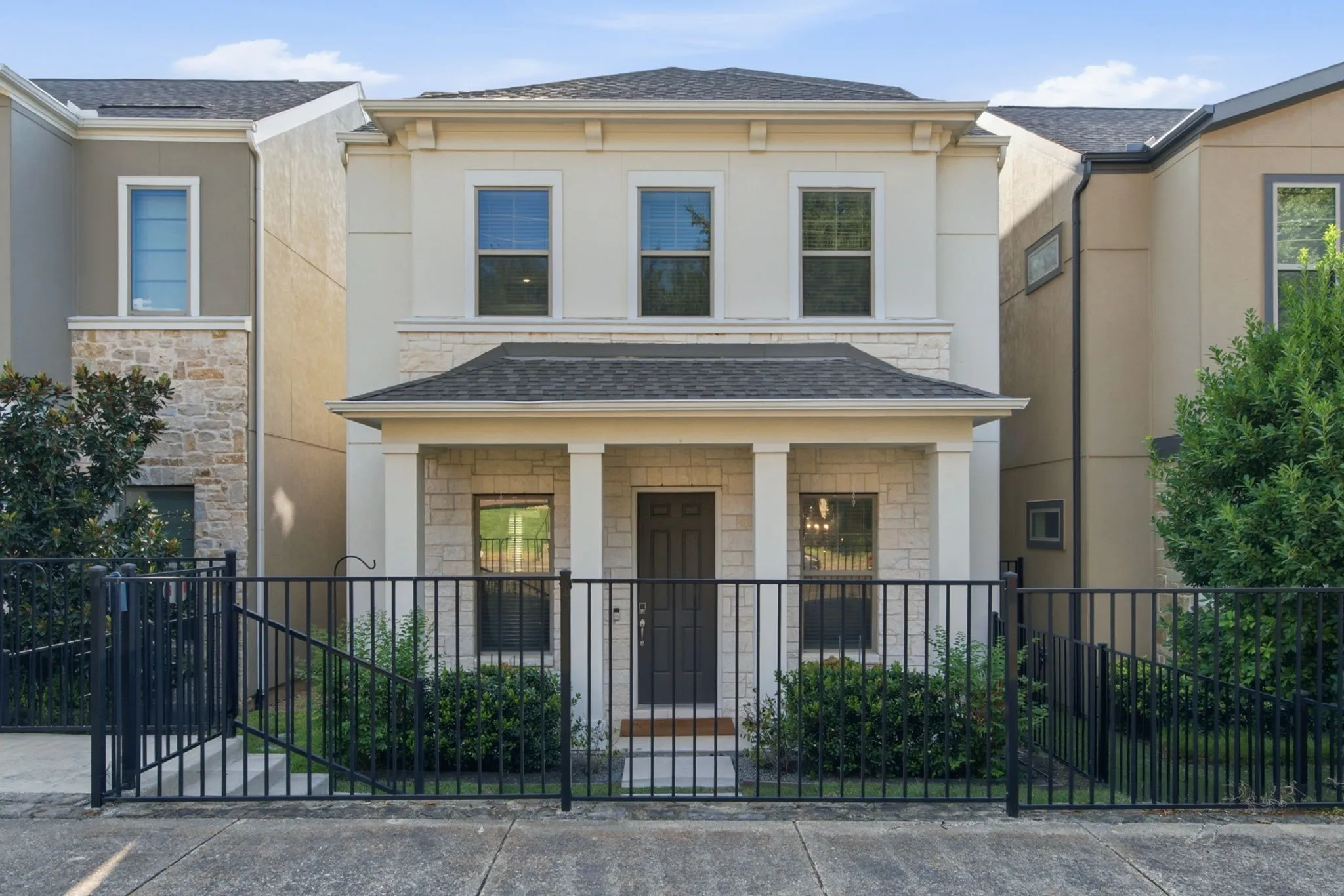 Two-story facade with stone and stucco exterior, covered front porch with columns, dark roof shingles, and black metal fencing