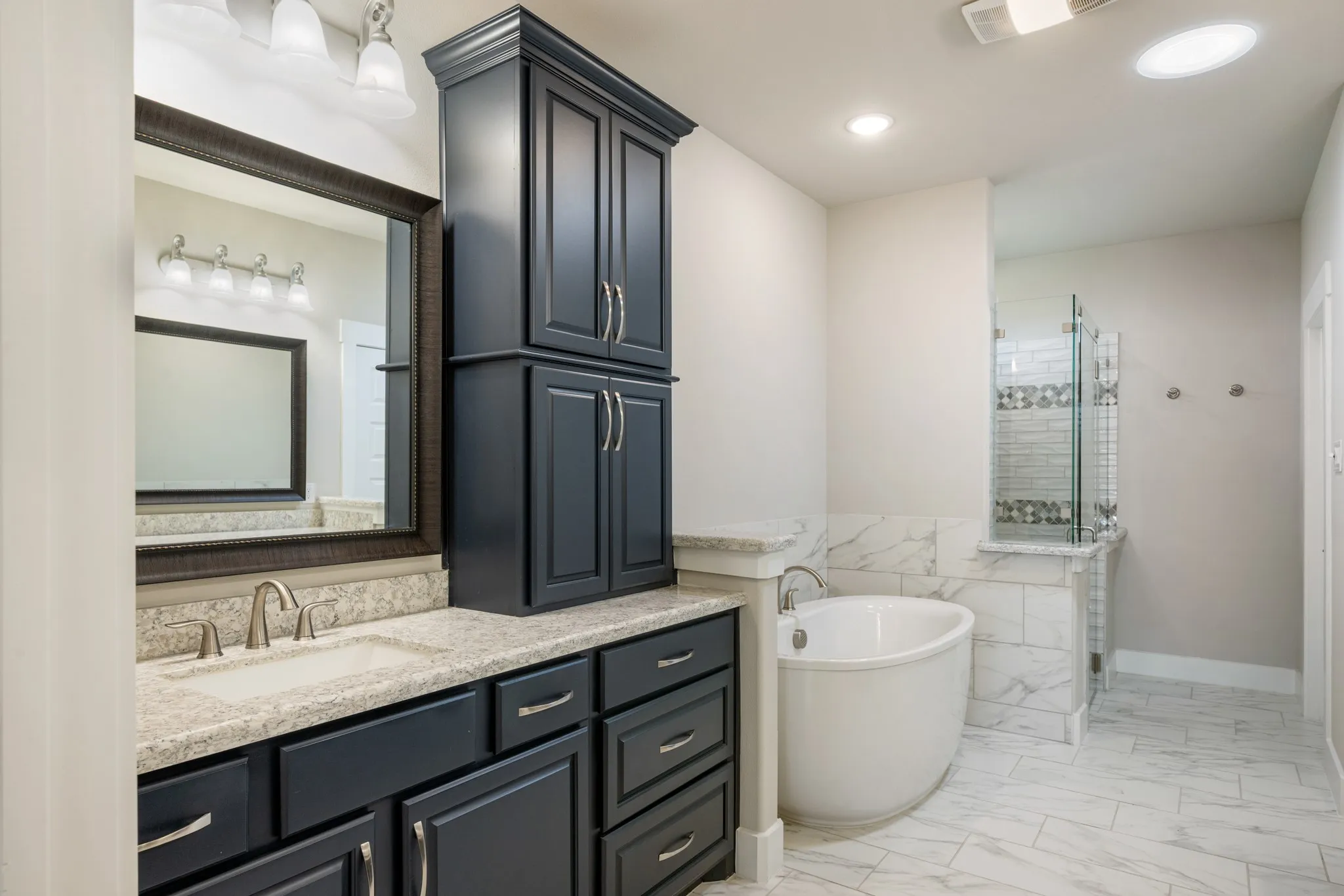 Spa-inspired primary bathroom featuring dual vanities with granite countertops, abundant cabinetry, a freestanding soaking tub, and a spacious walk-in shower with glass enclosure and decorative tile accents. Light, neutral finishes create a bright and inviting retreat.