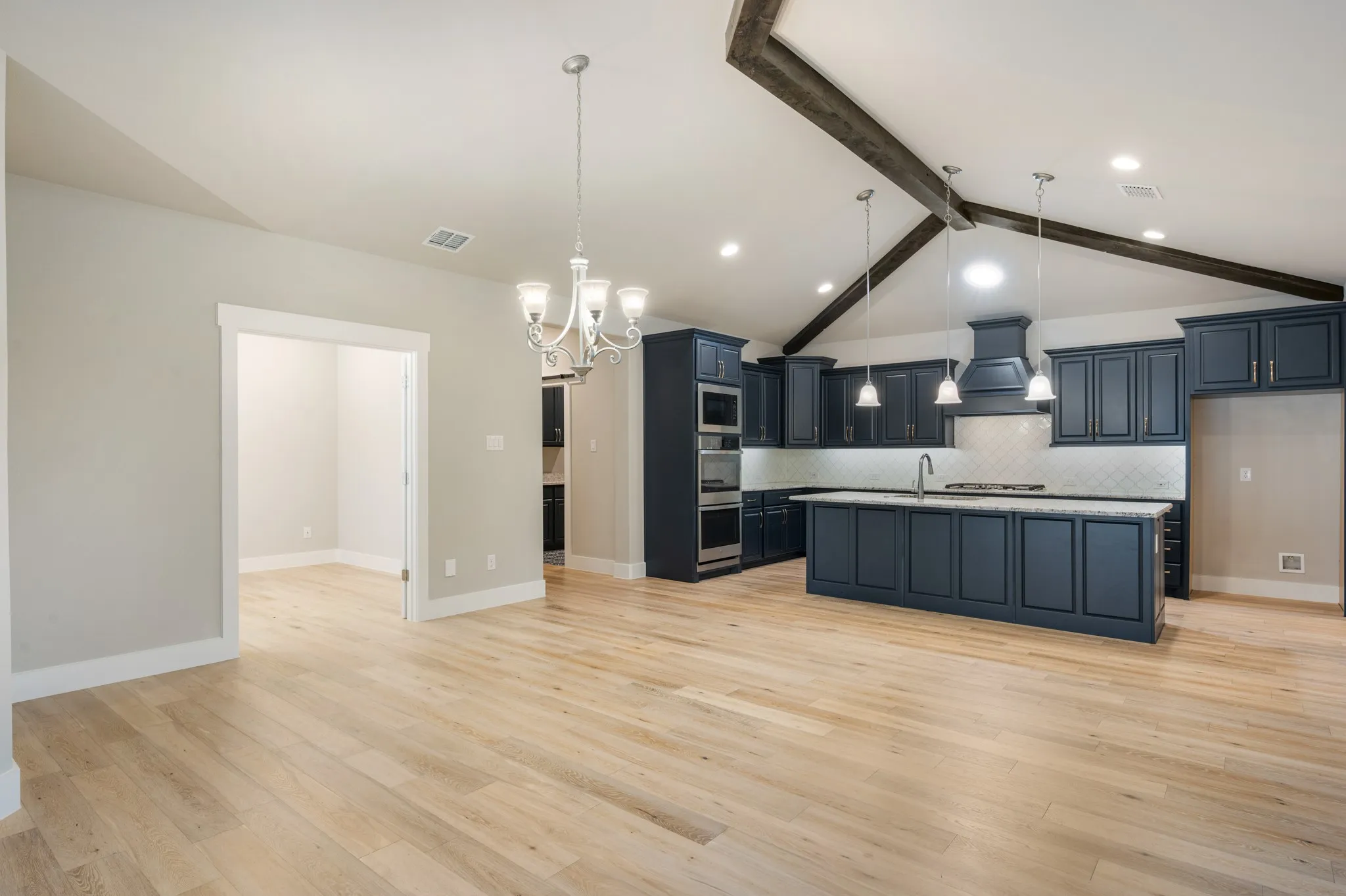 Bright, open-concept living space showcases beautiful wood-look flooring, vaulted ceilings with exposed wood beam accents, and an inviting flow into the spacious kitchen. The large center island, dark cabinetry, and stylish pendant lighting create a seamless space ideal for everyday living and entertaining.