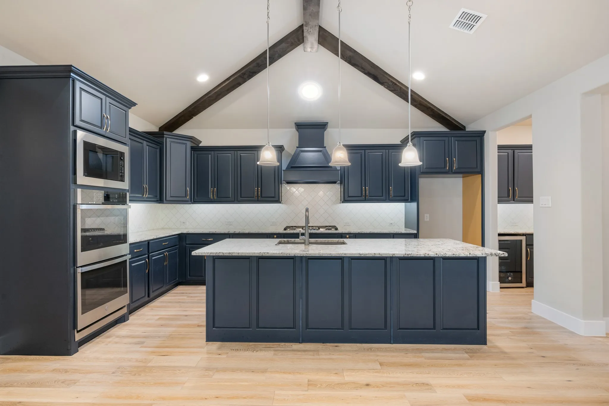 Spacious gourmet kitchen featuring a large center island with granite countertops, abundant custom cabinetry, stainless steel appliances, decorative tile backsplash, and a statement range hood. Vaulted ceiling with exposed wood beam accents adds character and an open, inviting feel.