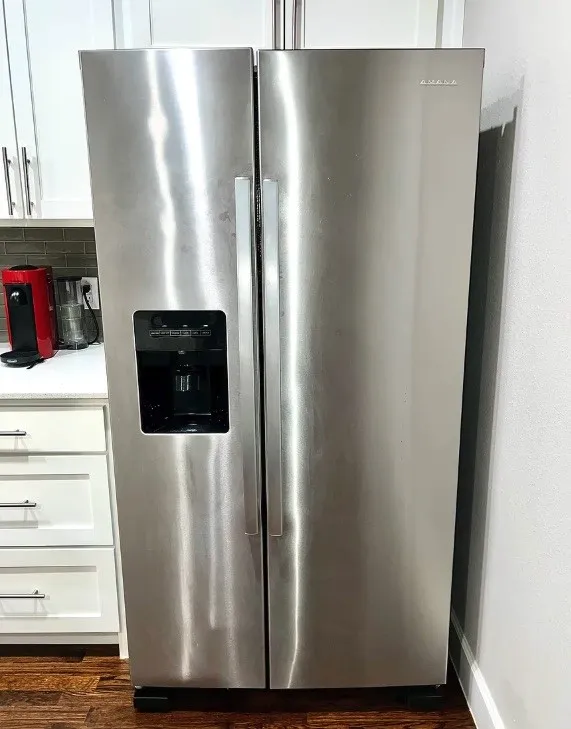 Stainless steel side-by-side refrigerator with integrated ice and water dispenser, set within a kitchen featuring white shaker cabinetry, white countertops, and a subway tile backsplash