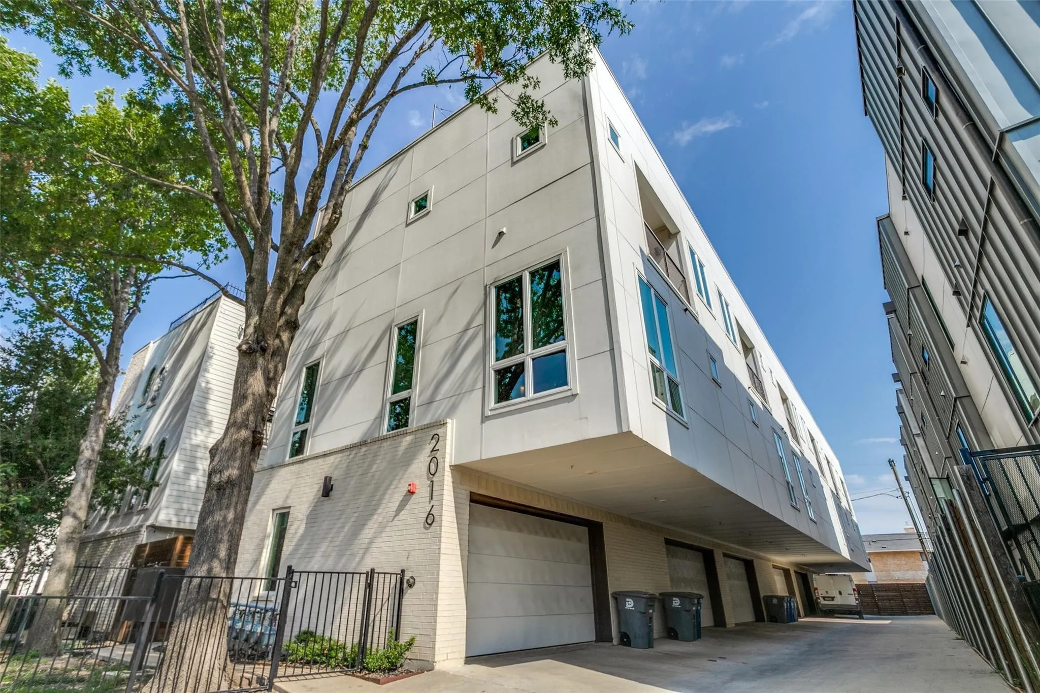 Modern exterior design with a white panel facade, ground-level garage access, tall vertical windows, a small upper-level balcony, and a black wrought iron fence